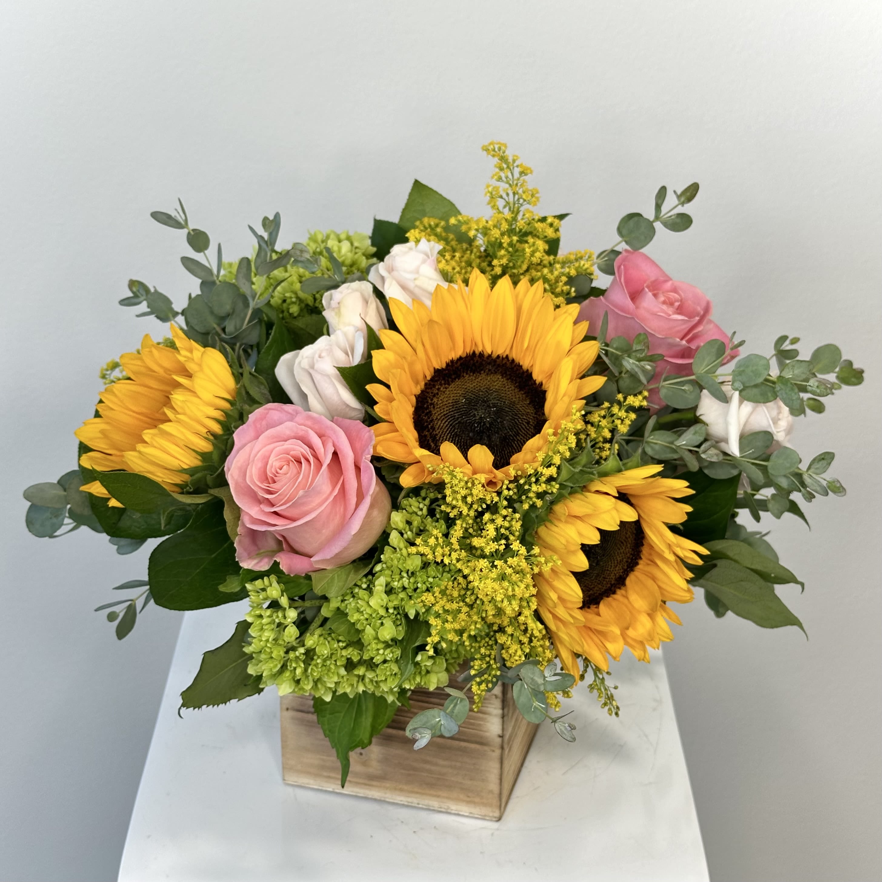 Sweet Sunflowers  - Sweet Sunflowers Flower Arrangement – Cheerful Sunflower Bouquet with Pink Roses &amp; Hydrangeas Illuminate any space with the "Sweet Sunflowers" arrangement, a delightful fusion of golden sunflowers and soft colored blooms. This handcrafted bouquet radiates warmth and joy, featuring bold sunflowers as the centerpiece, surrounded by romantic pink roses, airy white spray roses, lush green hydrangeas, vibrant solidago, and lemon leaves. Perfect for celebrating life’s sweetest moments!  Why Choose Sweet Sunflowers? Sunflowers: Iconic symbols of happiness, loyalty, and adoration, their large, sunny faces instantly uplift moods. Pink Roses: Represent gratitude, appreciation, and gentle affection, adding a tender touch. White Spray Roses: Delicate clusters bring elegance and purity, ideal for weddings or sympathy gestures. Green Hydrangeas: Voluminous and textured, they evoke serenity and abundance. Solidago: Golden-yellow plumes enhance brightness and fill the arrangement with natural fullness. Lemon Leaves: Shiny foliage provides a zesty green backdrop.  This versatile arrangement, nestled in a cedar wood box, suits birthdays, anniversaries, thank you gifts, or get well wishes. Sunflower bouquet, pink rose arrangement, sweet sunflowers delivery, solidago flowers, cheerful sunflower roses, same-day flower delivery, birthday sunflower bouquet, anniversary flowers, get well sunflowers. Order "Sweet Sunflowers" today and spread sunshine! Available for same-day delivery.