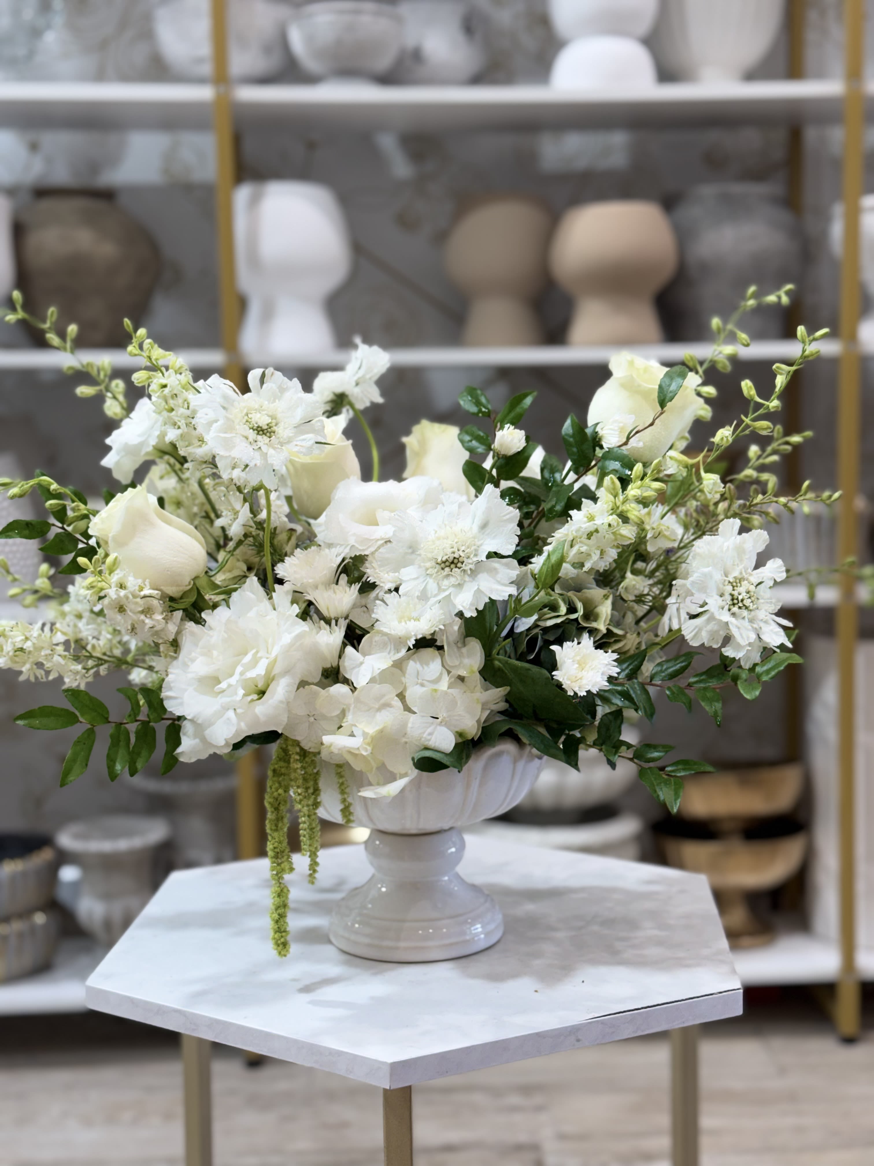 Blooms with love  - An airy cascade of white blooms arranged on a white pedestal vase creates a serene, elegant display that says "I'm thinking of you" with quiet confidence - perfect as an anniversary bouquet, a sympathy arrangement, or a refined birthday or get-well gift. The mostly white palette with touches of glossy green foliage brings a calm, timeless look that also reads romantic enough for love and romance or celebratory for Mother's Day and congratulations.  This flower arrangement blends delphinium, hydrangeas, lilies, garden roses, snapdragons, roses, daisies and complementary seasonal blooms into a broad, rounded design. Rounded hydrangea-like clusters sit beside delicate rose buds and daisy-like flowers, with tall snapdragons and delphinium adding vertical movement; all are gathered in a white pedestal vase that lifts the composition and suits a mantel, bedside table, or reception area. The overall size is generous and spreads broadly to either side for a full tabletop presence.  Choose this arrangement to comfort someone who is grieving, to celebrate a milestone, or to brighten recovery - the recipient will feel seen and cared for by its refined simplicity. Standard care: keep fresh water in the vase, trim stems every few days, and place away from direct heat or drafts to prolong bloom life.
