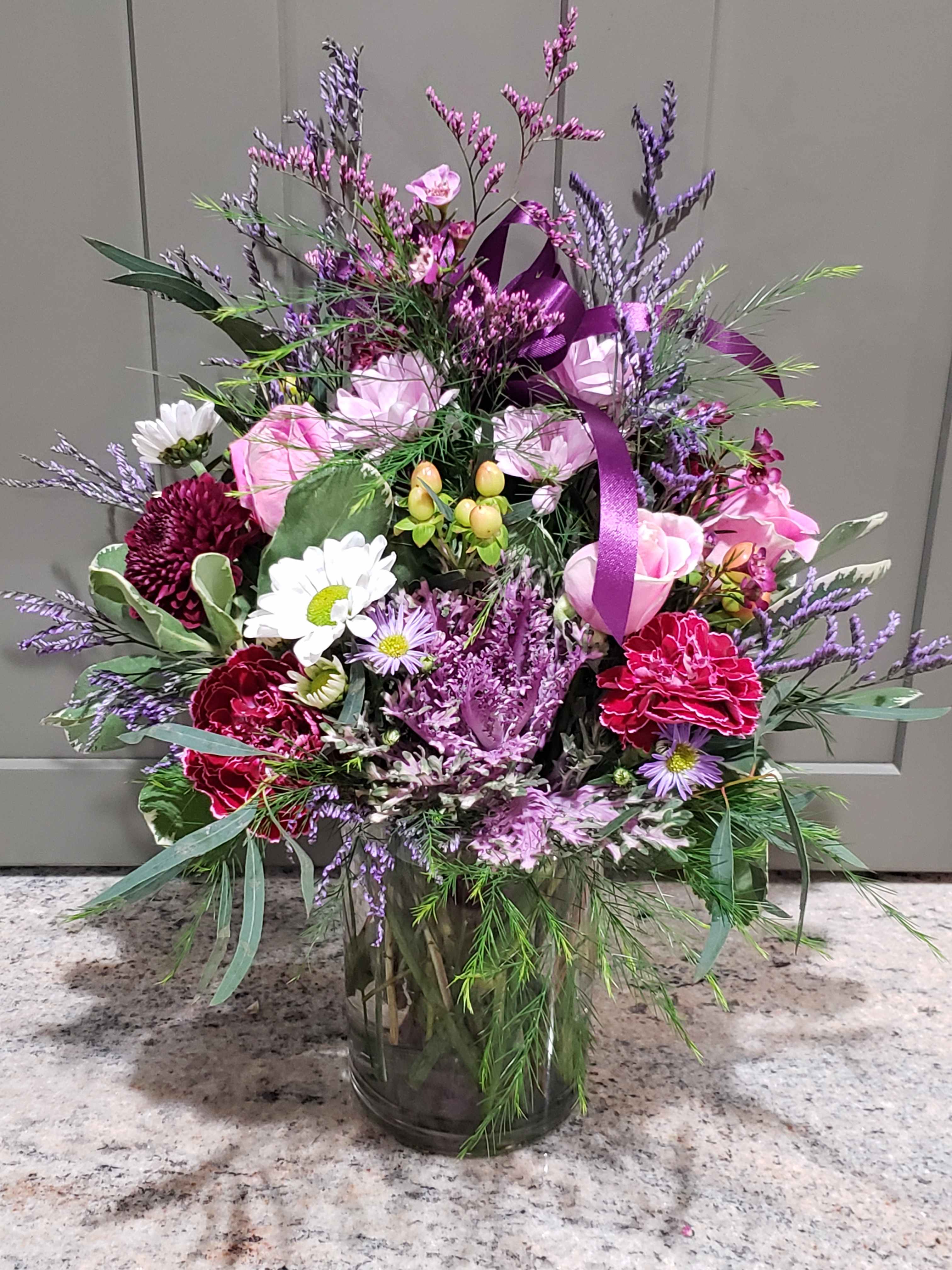 Tall mixed bouquet of pink roses, white daisies, and purple blooms in a clear glass vase with a purple ribbon.