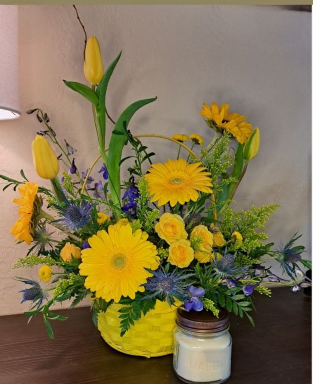 Sunshine Serenade - Bright yellow tulips, gerbera-style daisies and roses with blue-purple accents in a woven basket, garden-style arrangement with a glass candle for a cheerful, rustic gift. Can be customized in size for a larger price. 