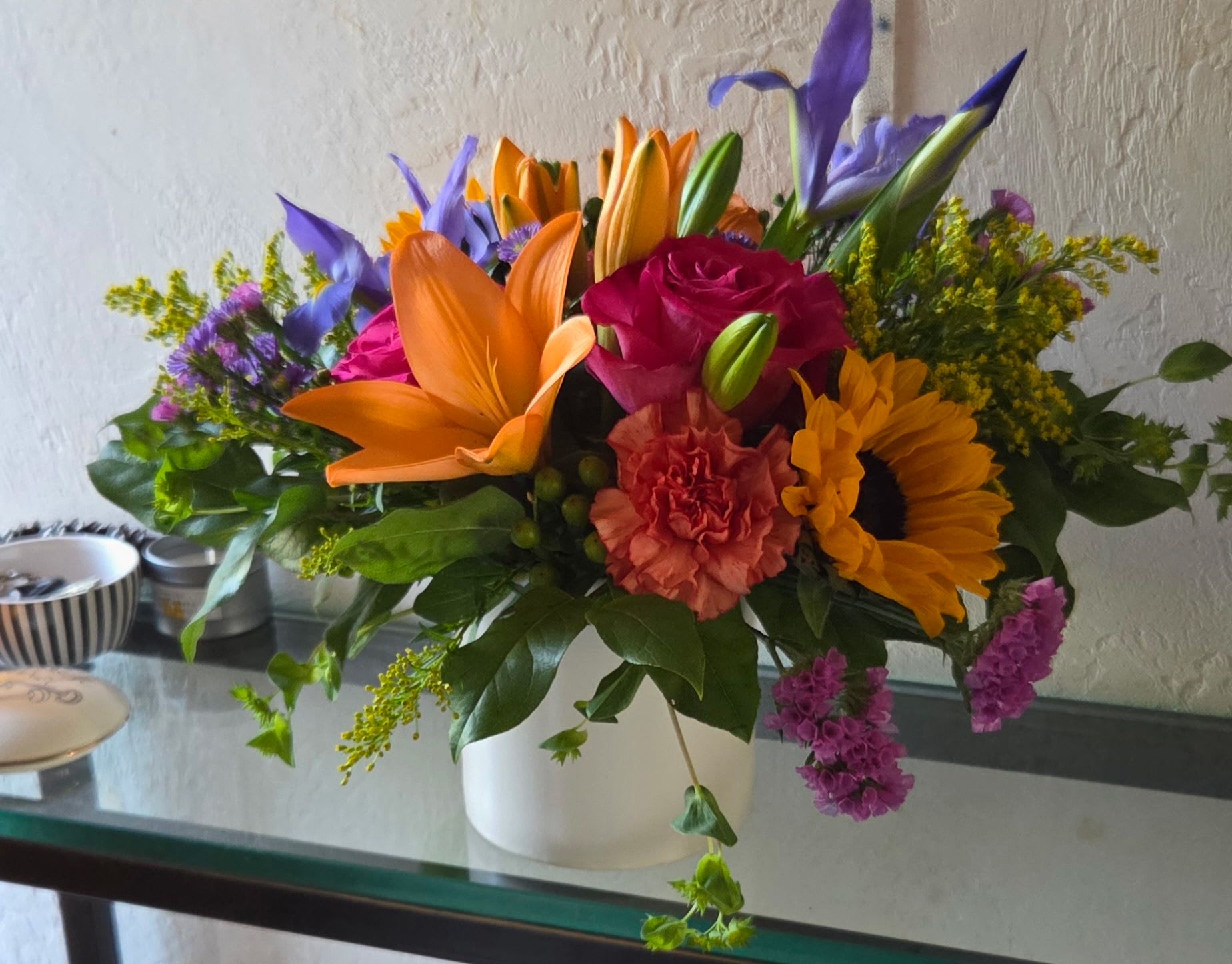 Sunset Symphony - White roses and mixed blooms in cool purples, blues and pinks, with bright orange lilies and sunflowers-layered, garden-style centerpiece finished with a blue ribbon or white ceramic vase. Can be adjusted for larger size for increased price. 