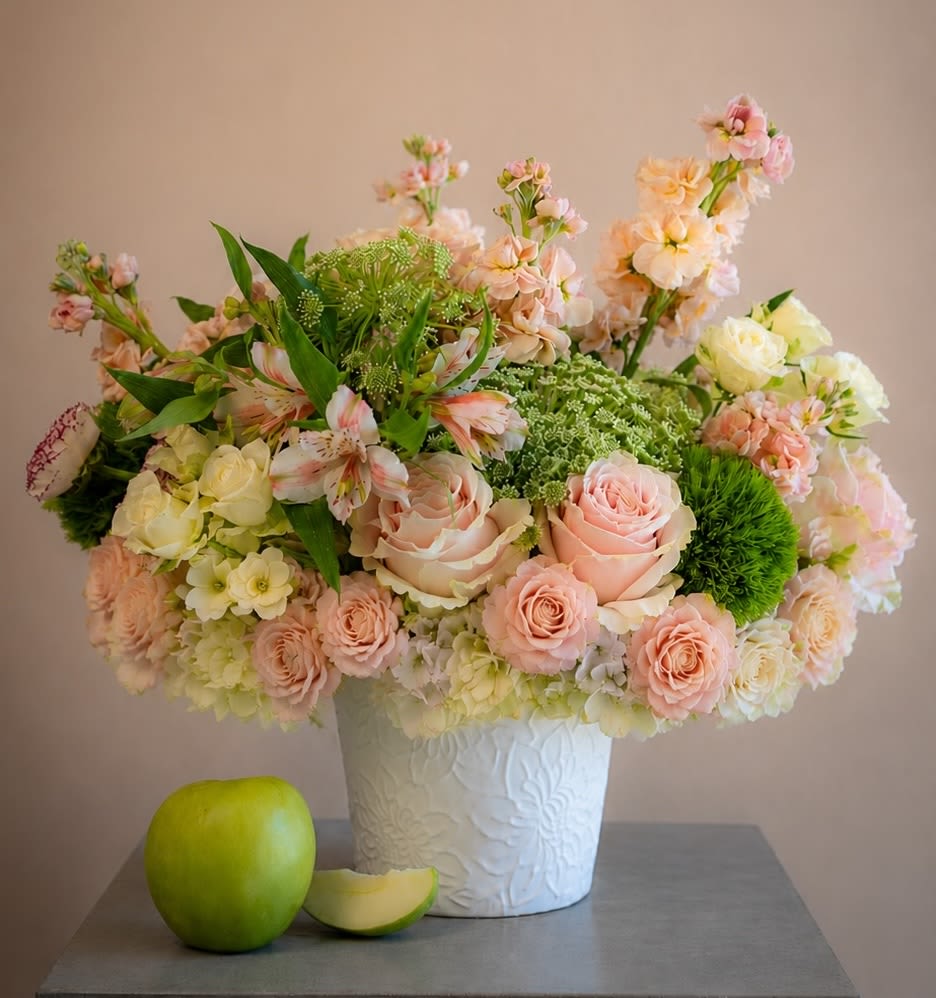 Rosa Prezioso (NEW) - A softly romantic centerpiece that makes someone feel delightfully cherished - Rosa Prezioso is a round floral arrangement ideal for a birthday bouquet, anniversary roses alternative, or an elegant congratulations gift. Blush and cream roses take center stage alongside pale hydrangeas and soft alstroemeria, creating a gentle pink-and-white palette that reads timeless and refined while bright green accents bring a fresh, springlike lift.  The composition is full and rounded, built with airy green filler flowers that add light texture and contrast. It arrives in a white textured (embossed) vase that enhances the arrangement's soft elegance; a green apple and a sliced apple are styled in front as a tasteful prop to emphasize the crisp green highlights. Colors include pink, white, green, and touches of yellow, and the mix of roses, hydrangeas, alstroemeria and seasonal filler gives the bouquet both volume and delicate scent.  Perfect for the person you want to make feel truly seen-whether celebrating a milestone, saying congratulations, or sending a thoughtful thinking-of-you gift-Rosa Prezioso suits formal tables and intimate rooms alike. For best enjoyment, keep flowers in fresh water and out of direct sunlight; available in a single rounded size as shown.