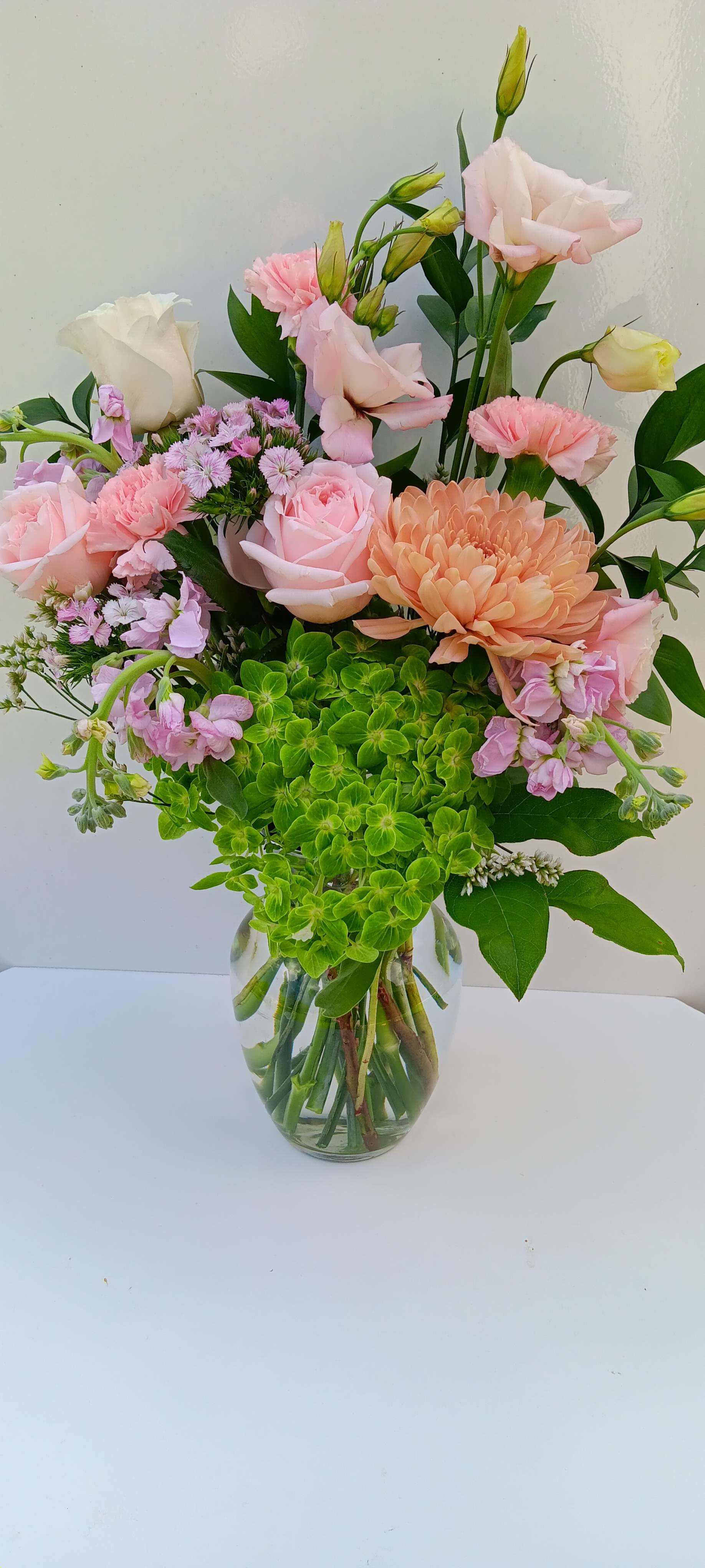 Tall glass vase of pink roses, carnations, lisianthus, peach mum, and green hydrangea.