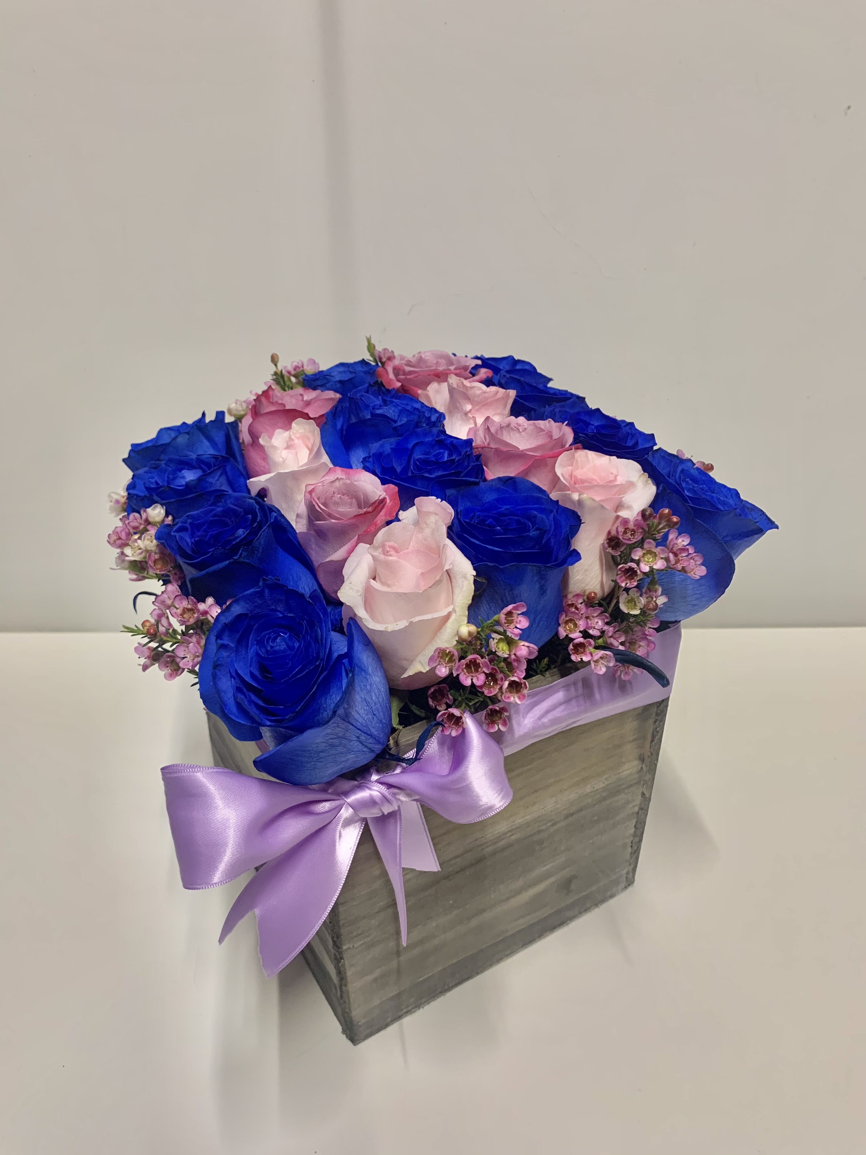 Square wooden box of bright blue and light pink roses with tiny pink blooms and a lavender bow.