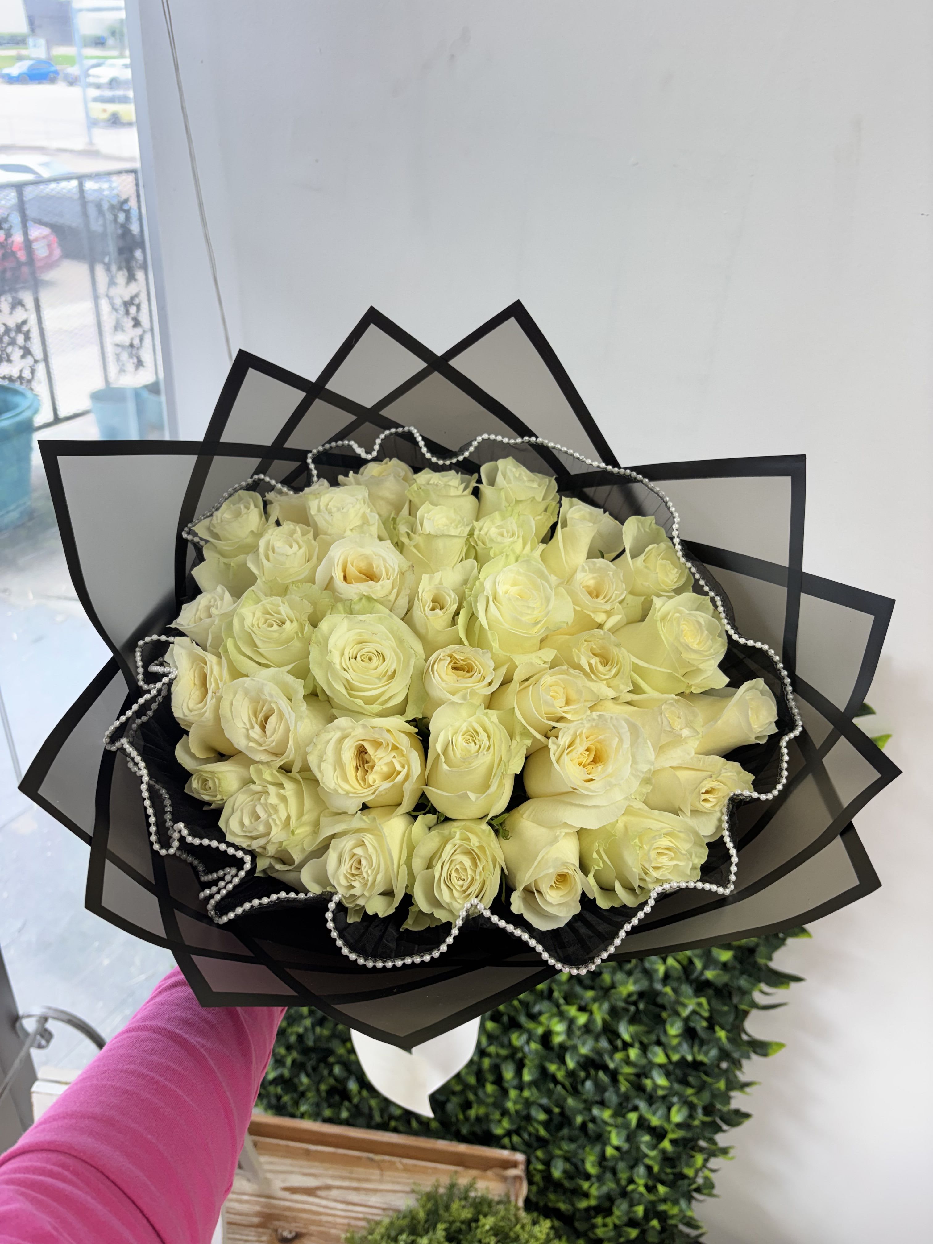 2 Dozen White Roses - Pale yellow roses gathered in a dense, rounded bouquet create a warm, romantic statement perfect for love and romance or a simple "just because" surprise. The compact, full shape lets the soft petals read like a single glowing bloom-an immediate, intimate impression that will make the person you're thinking of feel cherished.  Two dozen roses are tightly packed and wrapped in layered black translucent paper with a scalloped pearl trim for refined contrast, finished with a white ribbon for a polished, gift-ready look. The color palette of soft yellow blooms against black wrap and pearl detail reads elegant and timeless, ideal for anniversaries, romantic gestures, or lifting someone's spirits any day of the year.