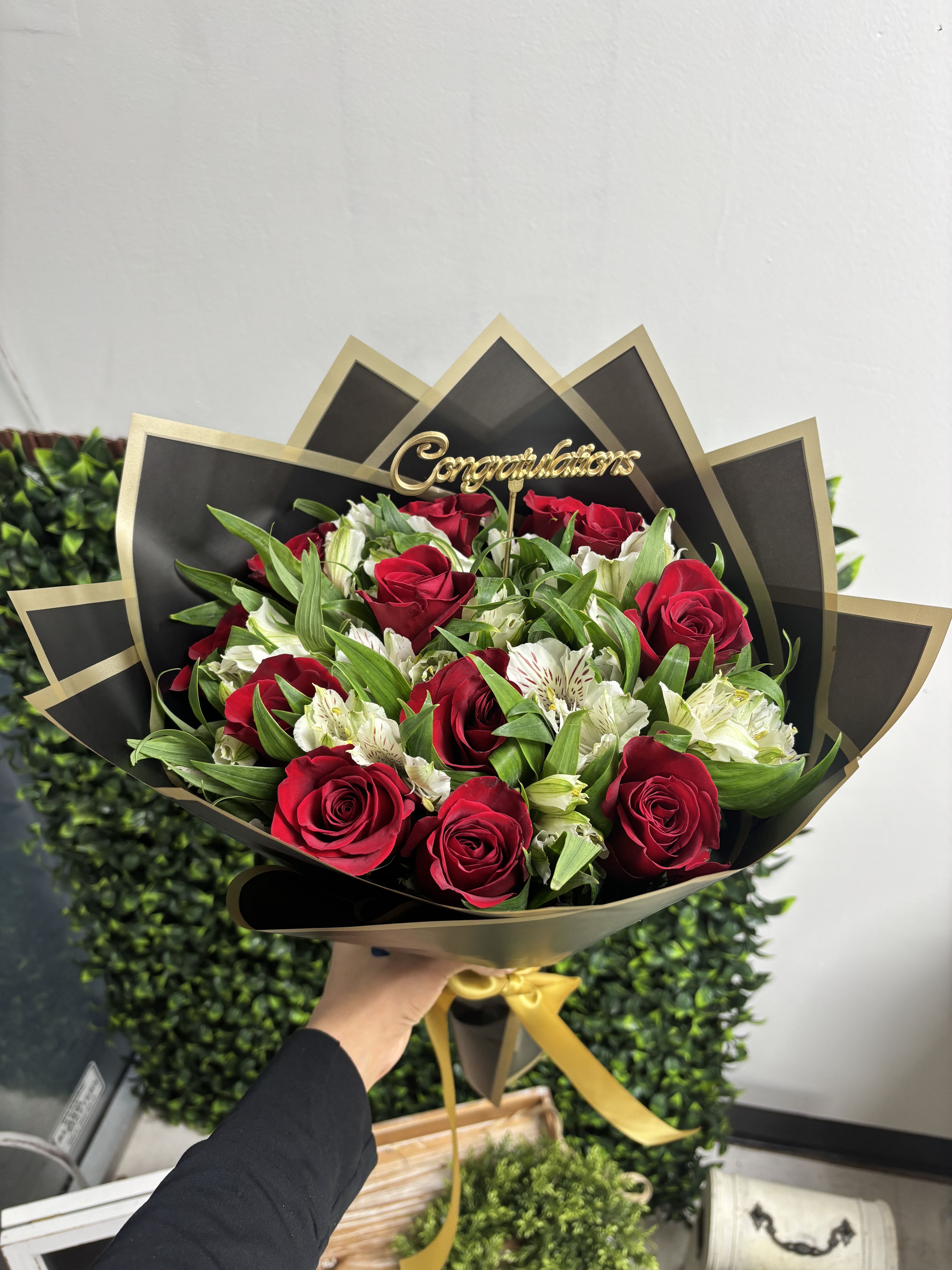 Congratulations Bouquet  - Tightly clustered red roses paired with crisp white alstroemeria create a bold, celebratory statement-perfect as a congratulations bouquet for graduations, promotions, or any milestone worth honoring. The rich red and bright white pop against layered black wrap, while the gold ribbon and a raised gold "Congratulations" topper make the occasion unmistakable and add a refined, gift-ready flourish.  This arrangement combines classic romance and formal polish: velvety red roses, delicate white alstroemeria, lush green foliage, and a dramatic dark paper wrap edged in gold. The bouquet is finished with a gold ribbon at the base and the topper set among the blooms for height and focus. The compact, tightly arranged composition keeps the flowers feeling abundant and elegant without being fussy.  Give this to the person you want to spotlight-the kind of bouquet that makes someone feel celebrated and seen, whether marking a new job, a degree, or a big personal win. Designed to arrive ready to display, standard care is to trim stems and place in fresh water; for longest life, keep away from direct heat and change the water every other day.
