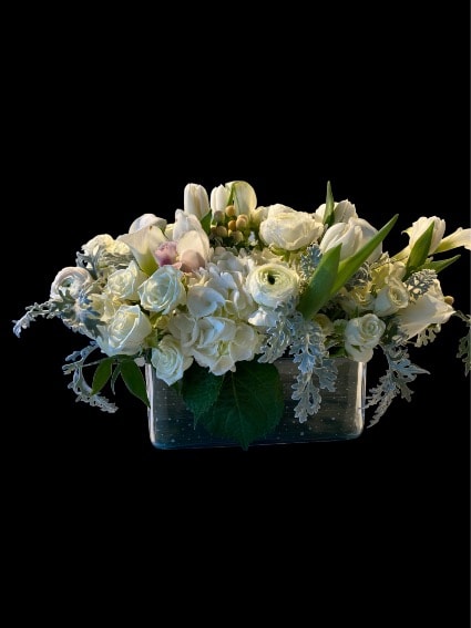 SOFT AND WHITE  - A low, rectangular centerpiece of pure white blooms creates an instant atmosphere of calm-perfect as an anniversary bouquet or a thoughtful way to say "thinking of you." This horizontal flower arrangement combines hydrangeas, creamy roses, tulips, calla lilies and ranunculus to form a continuous, mounded line across a clear rectangular glass vase, offering a refined, contemporary look that translates well to get well wishes, sympathy and funeral settings, or a simple thank you.  Closely clustered blossoms produce a soft, cloud-like texture, while silver-toned foliage and a few fresh green leaves provide subtle contrast that keeps the palette serene and sophisticated. The compact, elongated profile makes it ideal for a mantel, dining table runner, or bedside table where a low silhouette is preferred.  Designed to make the recipient feel quietly comforted and seen, this arrangement reads as both elegant and approachable-appropriate for formal occasions like weddings or for intimate gestures of care. Arrives ready to display in its glass vase; keep in a cool spot away from direct sun and change the water regularly to extend freshness.