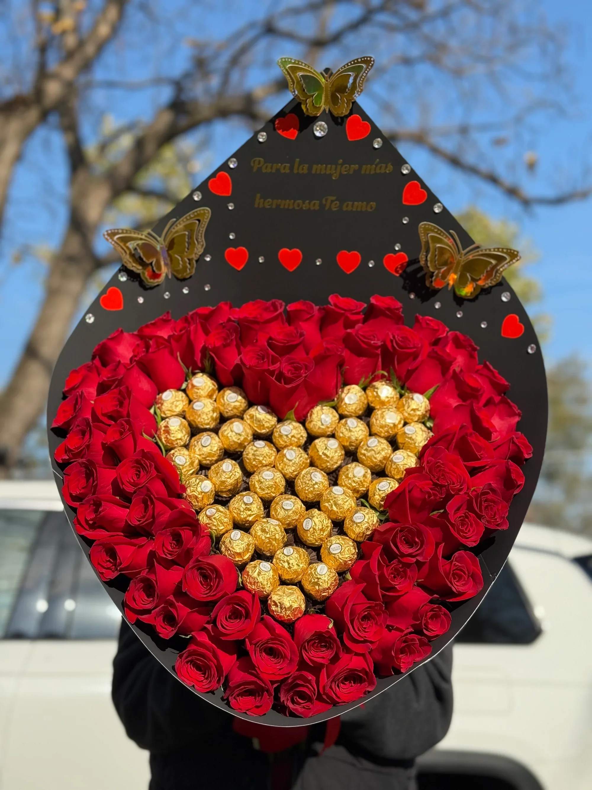Ramo con chocolates - ️ Ramo Corazón de Rosas con Chocolates – Edición Romántica   Declara tu amor de una manera inolvidable con este impresionante ramo en forma de corazón. Elaborado con hermosas rosas rojas frescas que rodean un delicado centro de chocolates premium, formando un corazón dorado que simboliza amor y dulzura.  Su elegante envoltura en tono negro, decorada con mariposas doradas y pequeños corazones, incluye el mensaje: “Para la mujer más hermosa, Te amo”, convirtiéndolo en el regalo perfecto para expresar tus sentimientos más profundos.   Ideal para:  San Valentín Aniversarios Cumpleaños Declaraciones de amor Sorpresas románticas   Incluye:  Rosas rojas frescas de alta calidad Centro de chocolates tipo Ferrero Diseño en forma de corazón Envoltura premium decorada Presentación lista para regalar   Un detalle elegante, dulce y lleno de pasión que conquistará su corazón desde el primer momento. ️  Haz tu pedido hoy y sorprende con un regalo que combina flores y chocolates en una presentación espectacular.