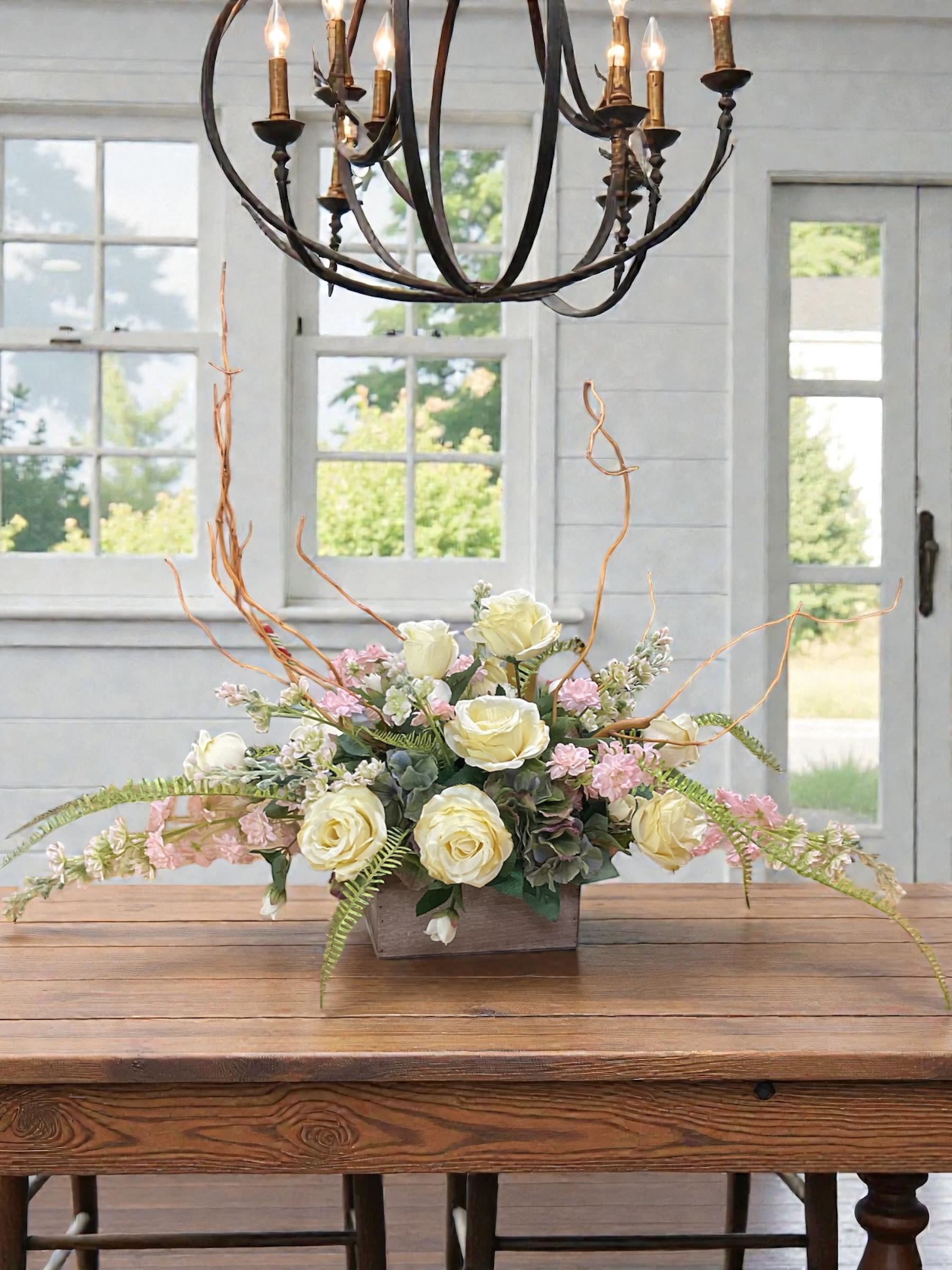 Countryside Grace Silk Arrangement - Countryside Grace is a beautifully styled silk arrangement designed to bring warmth, elegance, and timeless charm to any home or office. Featuring creamy ivory roses, soft blush blooms, natural-looking greenery, and graceful willow branches, this design blends rustic character with refined French-country style.  The low, elongated shape makes it ideal for dining tables, coffee tables, kitchen islands, buffets, mantels, entry tables, and reception areas. Flowing lines and carefully placed branches create movement and visual interest without overwhelming the space, making it a versatile centerpiece for everyday decorating.  Soft neutral tones allow Countryside Grace to complement a wide variety of décor styles, including farmhouse, cottage, traditional, French country, and transitional interiors. The combination of ivory, blush, sage, and natural wood tones creates a relaxed, welcoming feel that works beautifully throughout the year.  This permanent botanical arrangement provides lasting beauty season after season. Whether displayed as a focal point on a dining table, accent piece on a console, or decorative touch in an office or reception area, Countryside Grace offers enduring style with the look of fresh-cut blooms.  A timeless silk floral arrangement designed to add beauty, texture, and graceful charm to any setting.