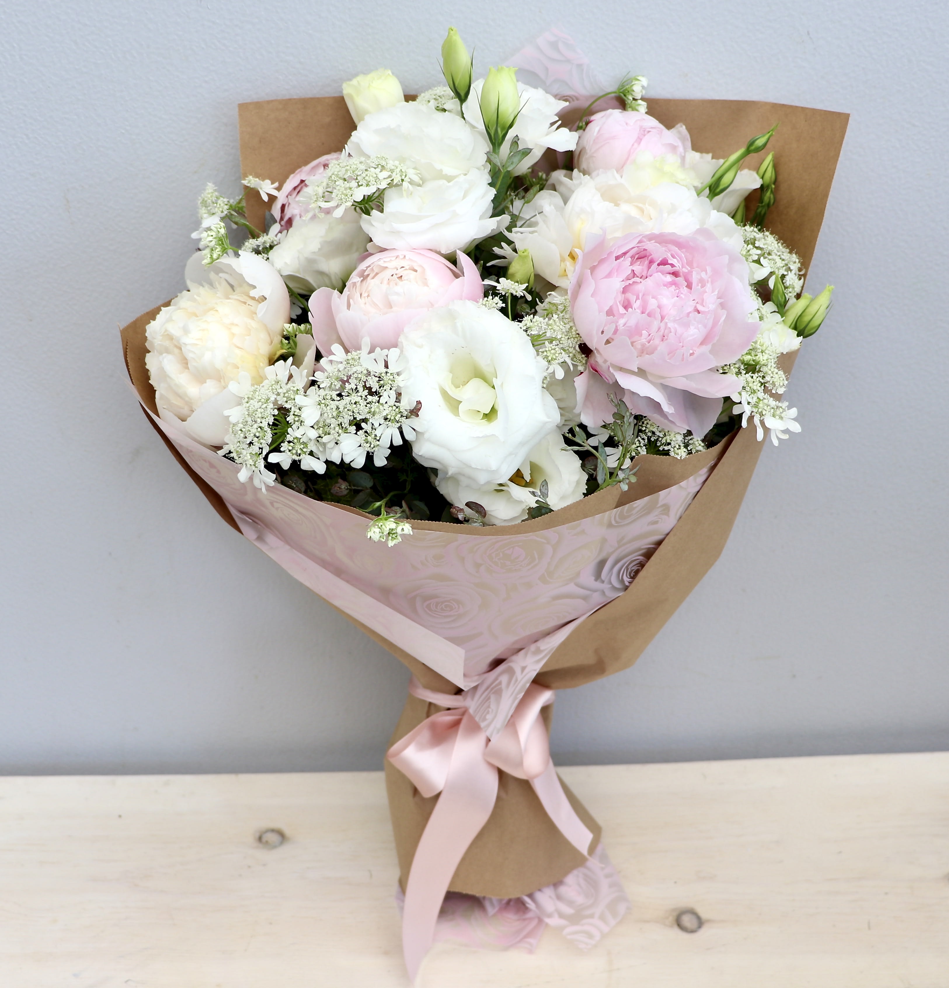 Hand-tied bouquet of white and pale pink peonies and lisianthus wrapped in kraft paper with a pink ribbon