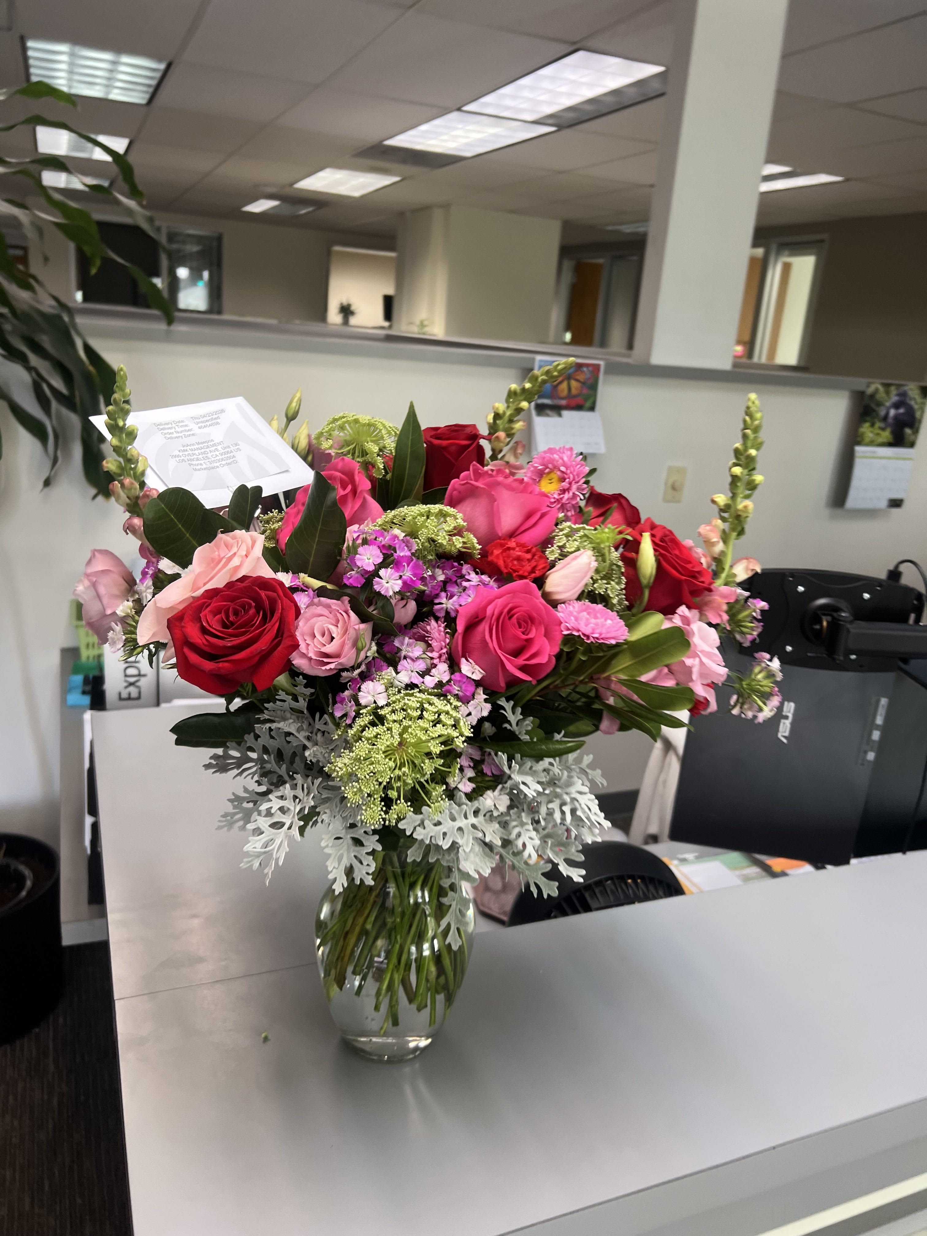 SEND A BIT OF JOY BOUQUET- ON SALE WAS $110 - A lush cluster of pink and red roses creates an immediate impression of romance and joy-ideal for a birthday bouquet, anniversary roses, or a heartfelt "thinking of you" gift. The romantic color mix of warm pinks, deep reds, crisp whites and fresh greens reads cheerful and polished, the kind of arrangement that makes the person you're sending it to feel genuinely celebrated.  This arrangement combines densely packed pink and red roses with smaller pink filler blooms, airy green accents, silvery foliage at the base, and delicate mat-form textures for depth, all presented in a clear glass vase with a thoughtful enclosure card. Finished for easy office or home display, it arrives ready to brighten a desk or dining table; water upon arrival and trim stems if desired to extend bloom life.