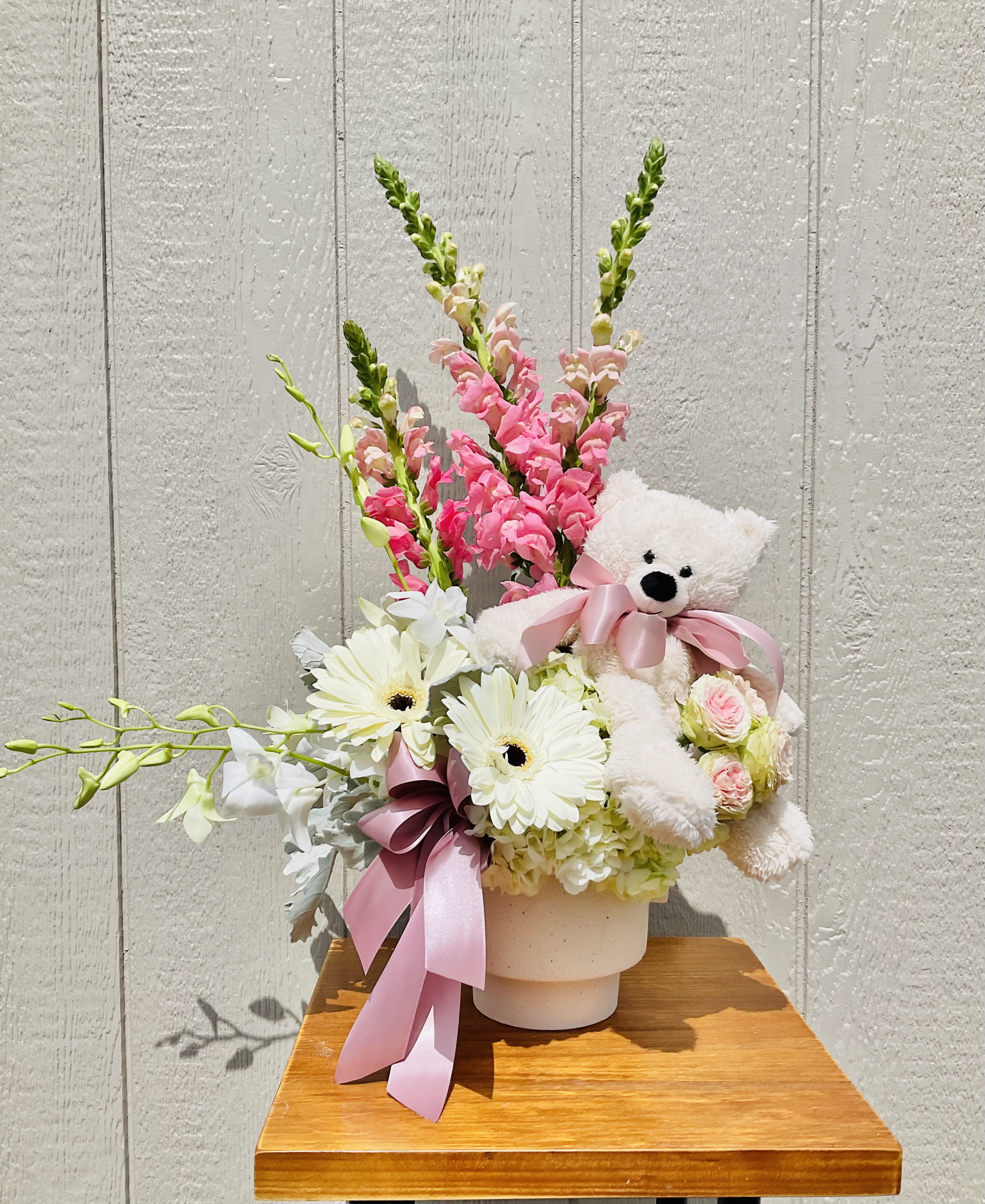 Hey Sweet Girl! - Upright pink snapdragons and crisp white gerbera daisies give this flower arrangement a sweet, celebratory silhouette-perfect as a birthday bouquet, a new baby gift, or a thoughtful "thinking of you" surprise. The mix of pale hydrangea blooms, spray roses and a few small pink roses creates a soft, layered palette of pinks, whites, and fresh green stems that reads gentle and cheerful to the person you're celebrating.  Arranged in a rounded white ceramic pot, the design pairs taller, moving stems with a fuller base of blooms for both height and plush texture. A plush white teddy bear wearing a pink satin ribbon is tucked into the side, adding a playful, gift-like touch that makes the recipient feel truly seen and comforted. Sized to sit on a bedside table or mantel, this arrangement arrives ready to display and enjoy-no fuss care: keep flowers in fresh water, away from direct heat, and they'll brighten the room for days.