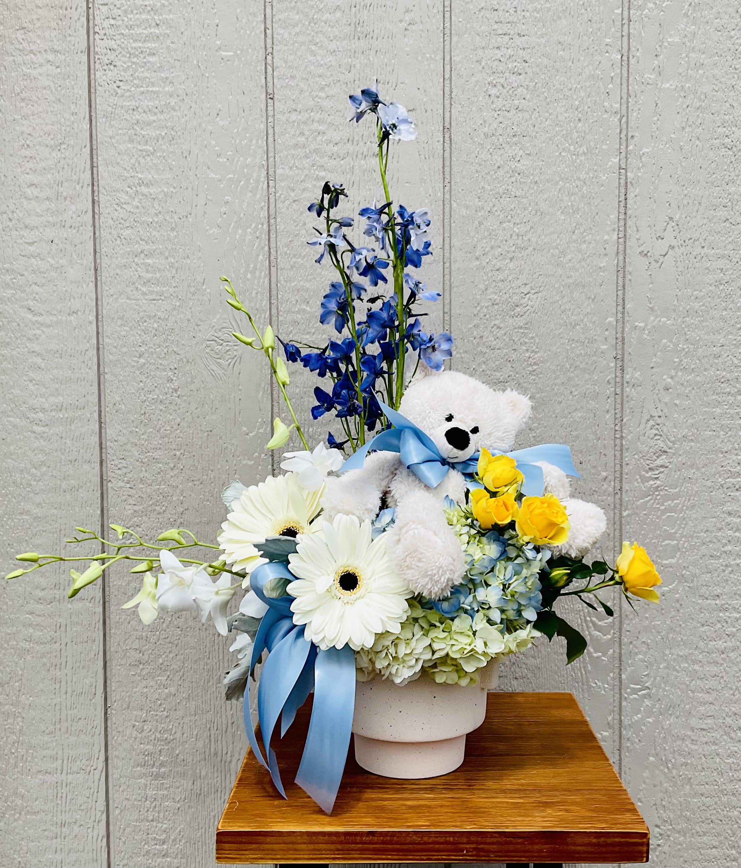 Hey Sweet Boy! - Bright, cheerful and perfectly playful, this flower arrangement is made for celebrating a new little one or sending birthday wishes-the kind of bouquet that makes the recipient feel seen and adored. White gerbera daisies, pale blue hydrangea and tall blue delphinium stems create soft, cloud-like volume while sunny yellow roses punctuate the design with a warm, joyful note; the overall palette of white, blue, yellow and green reads as both celebratory and tender for a baby boy or any happy occasion.  Arranged compactly in a ceramic pot, the composition is finished with a cuddly white teddy bear tied with a blue ribbon tucked into the blooms, turning the piece into an easy gift-and-keepsake in one. Textures range from the rounded hydrangea clusters to the sleek orchid stems and the cheerful daisy faces, giving visual interest from every angle. Sized to fit on a bedside table or mantel, it's a ready-made surprise for new parents, a cheerful birthday table accent, or an everyday pick-me-up that says "thinking of you" with sweetness and style.