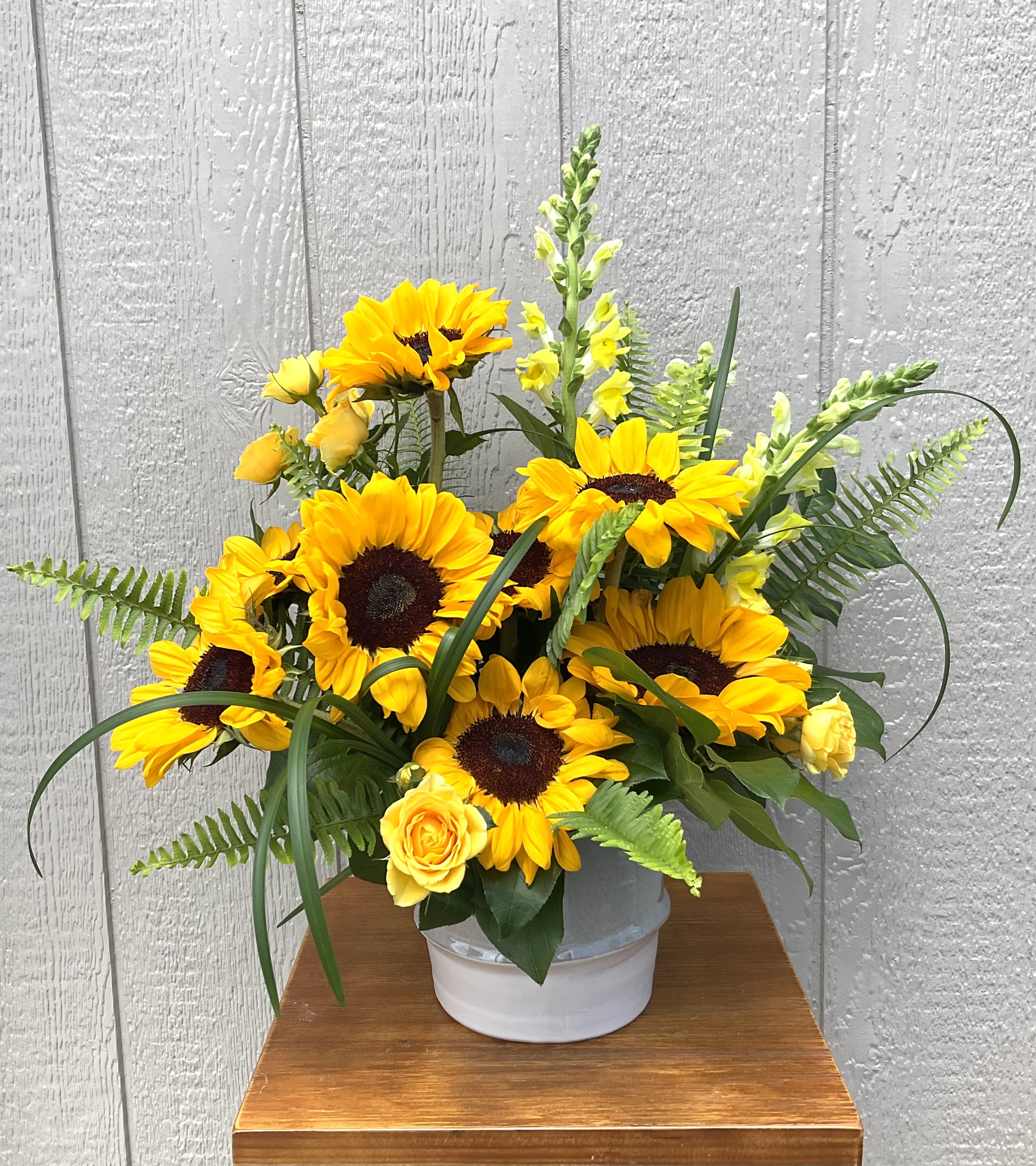 Twas’ All Yellow - A burst of sunlit cheer-this yellow sunflower arrangement is designed to make the person you're thinking of feel bright and celebrated. Perfect as a birthday bouquet, congratulations gift, or a just-because surprise, the strong yellow palette reads sunny and optimistic while the touches of green and white keep it fresh and natural.  Clusters of bold yellow sunflowers sit with smaller yellow roses and tall yellow snapdragon spikes for height and movement, all woven with arching foliage and fern fronds for a garden-style silhouette. The flowers arrive arranged in a clean white ceramic vase that anchors the color and keeps the presentation simple and modern. This is the kind of bouquet that brings instant warmth to a kitchen table, office desk, or bedside-no fuss care, just trim stems and fresh water to enjoy its cheerful bloom.