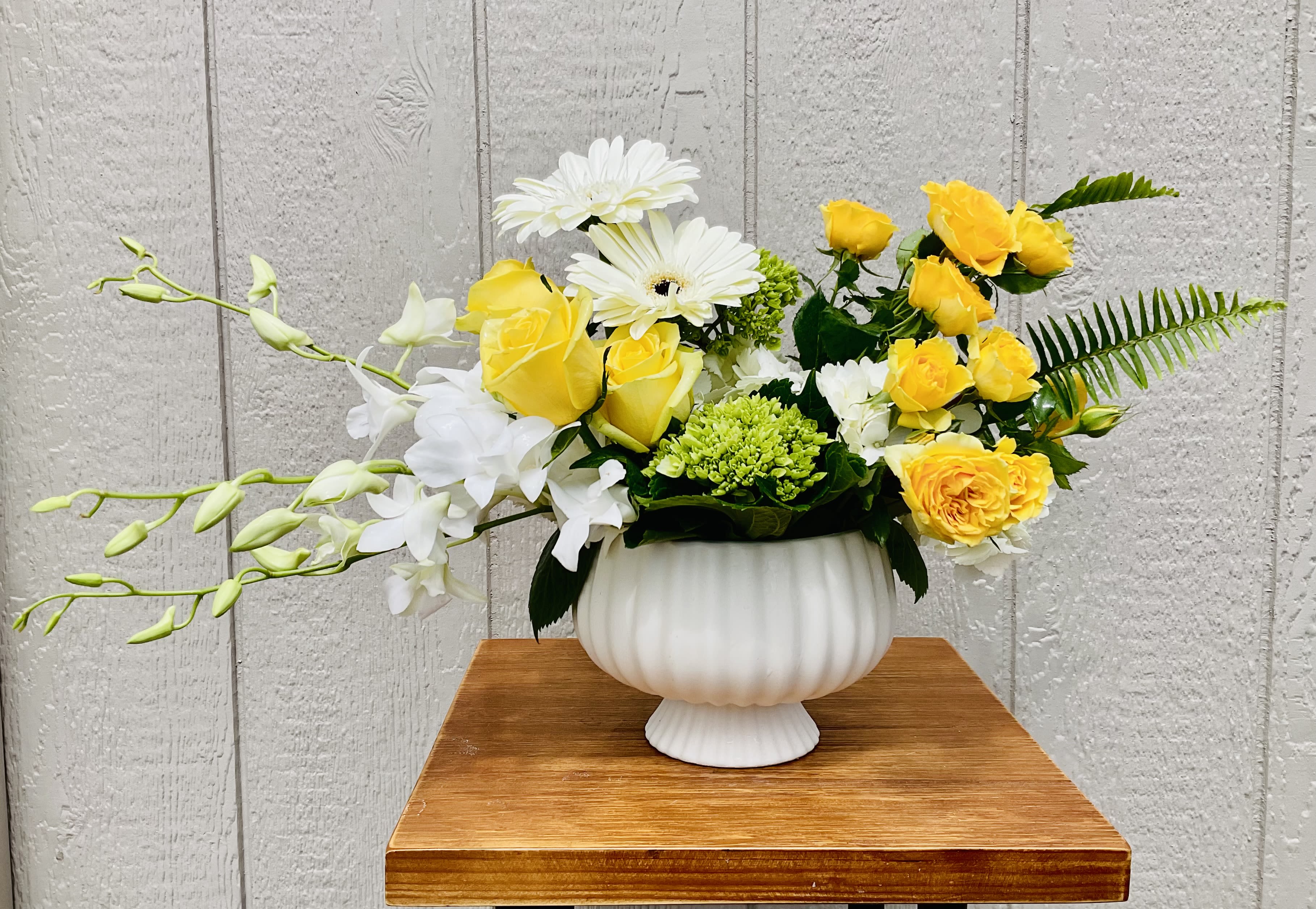 Sunny Day - Sunlit yellow roses and crisp white gerbera-like blooms meet elegant white orchid sprays in a cheerful bouquet designed for everyday celebration. This flower arrangement for birthdays, thank-you gestures, or springtime surprises gives the person you're thinking of a bright, uplifting gift that reads fresh and modern while still feeling warm and personal.  Arranged in a rounded white ceramic vase, the design mixes soft yellow, clean white, and fresh green foliage for a lively but balanced look. The composition is full and slightly asymmetrical - long orchid stems extend to one side while fern-like greenery reaches the other - creating an airy silhouette that feels both elegant and relaxed. Perfect for a kitchen counter, office desk, or bedside table, the Sunny Day arrangement is an easy way to make someone feel truly seen.