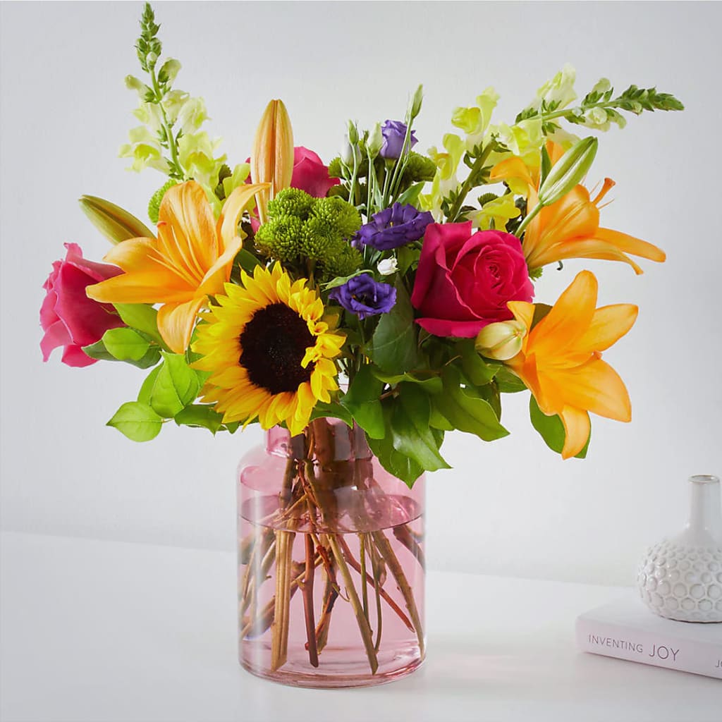 Colorful bouquet of roses, lilies, sunflowers, and purple blooms in a pink glass vase