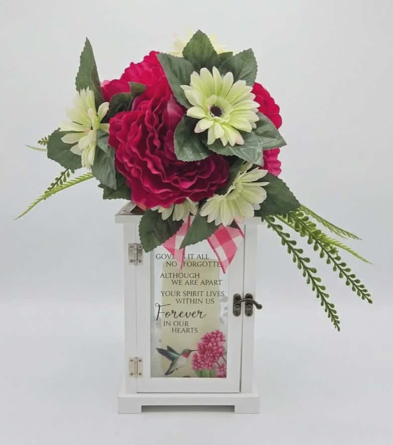 Forever In Our Hearts Lighted Lantern w/ Silk Floral - Bright pink rose-like blooms and pale white daisies crown a white memorial lantern in this sympathy arrangement designed to comfort and commemorate. As a funeral or remembrance piece, the Forever In Our Hearts lighted lantern with silk floral sends a gentle message of ongoing love-she'll see the sentiment every time the soft light glows and the pink ribbon adds a tender, personal touch.  The silk flowers are arranged with layered texture and long, trailing greenery that drapes gracefully to either side of the lantern, creating a balanced, evergreen look that requires no maintenance. The white wood lantern (6.25" W x 13.5" H x 6.25" D) features a stainless-steel copper-finish top, a built-in automatic timer (6 hours on / 18 hours off) and includes three AAA batteries. The lantern front is inscribed with the sympathy verse, making this an enduring, ready-to-display tribute suitable for funeral services, a graveside marker, or a heartfelt memorial at home.