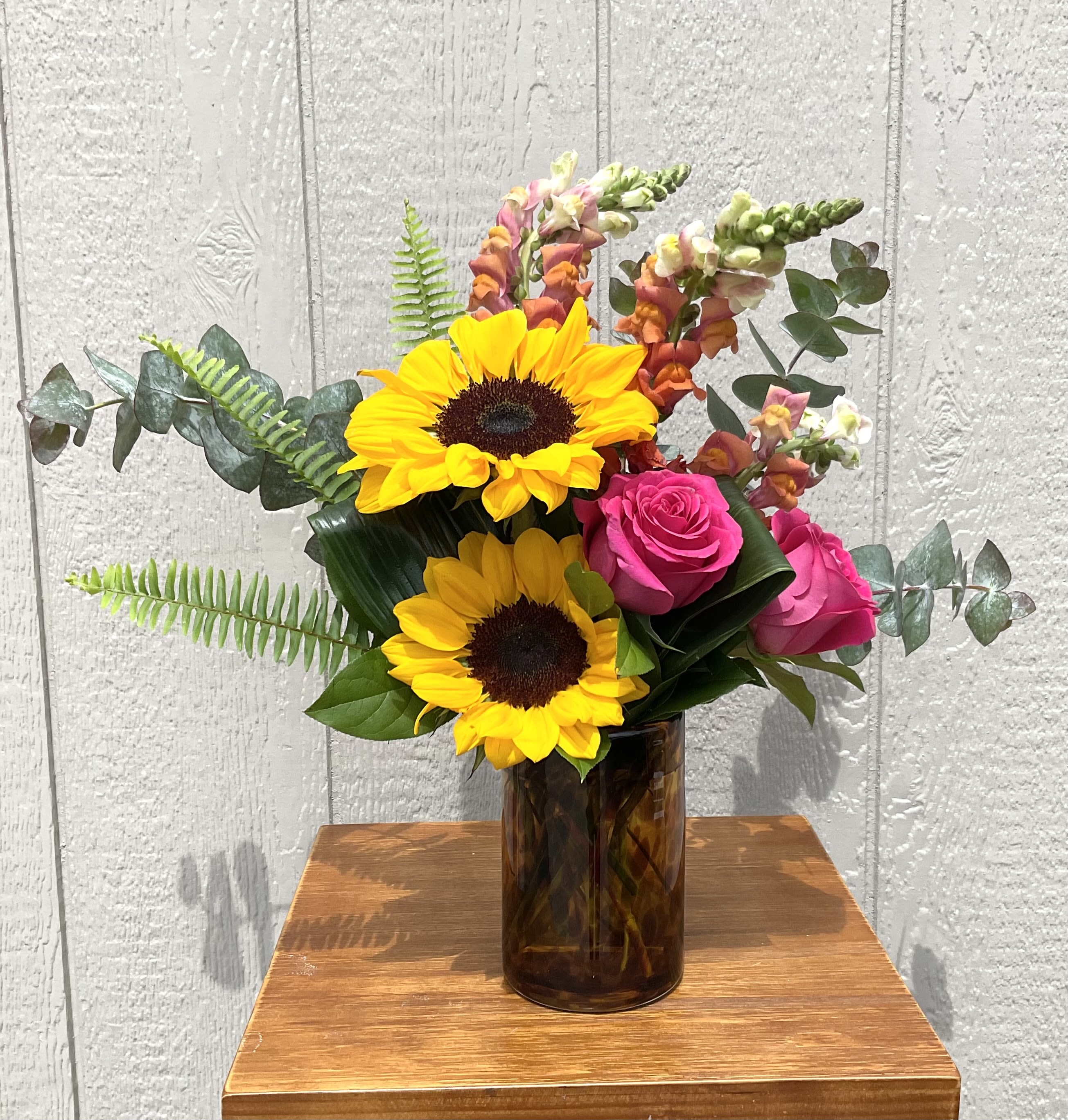 Jazzed. - Two bright yellow sunflowers take center stage in this cheerful flower arrangement, paired with vivid pink roses and tall snapdragon spikes in orange, pink, and cream-perfect as a birthday bouquet or everyday celebration arrangement. The warm palette of yellow, pink, orange and green creates an energetic, uplifting impression that will make the person you're thinking of feel noticed and celebrated.  The composition is compact but airy: sunflowers and roses clustered near the vase with snapdragons adding vertical movement, while eucalyptus and fern-like foliage extend outward for a loose, garden-style silhouette. Arranged in a dark amber brown glass vase, the piece has a modern yet earthy contrast that works well on a kitchen table, office desk, or entry console.  Ideal for birthdays, congratulations, or "just because" moments, this arrangement sends a bright, joyful message without being overly formal. Care is simple-fresh water in the vase and regular trimming of stems keeps the bouquet looking lively; choose delivery or local pickup to ensure the sunflowers and roses arrive at their freshest.