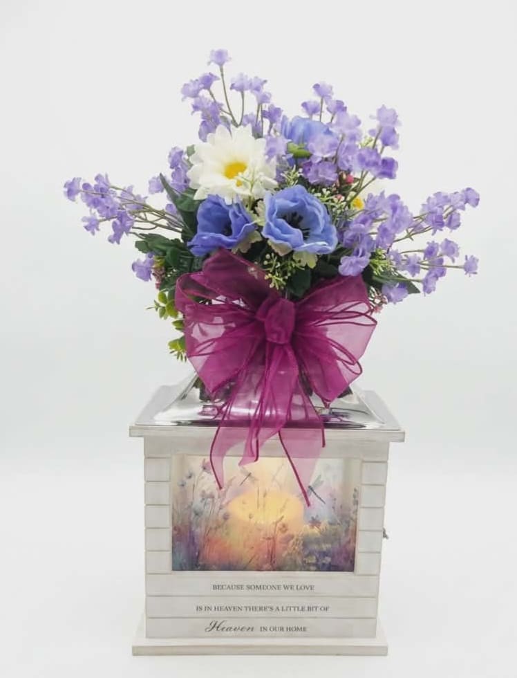 Heaven in Our Home Lighted Lantern  w/Silk Floral  - A cool-toned cluster of purple and lavender blooms rests atop a softly glowing decorative lantern-an elegant sympathy arrangement designed to comfort and commemorate. This flower arrangement for remembrance pairs gentle daisy accents and mixed silk blooms in shades of purple, blue, pink, and white with a printed memorial message on the lantern, making it a comforting choice for a funeral, memorial service, or a home remembrance display.  The white wooden lantern has stainless-steel-finish hardware, glass panels printed with a pastel landscape and the sentiment, "Because someone we love is in Heaven, there is a little bit of Heaven in our home." A loose cluster of purple and lavender silk flowers surrounds a white daisy-like bloom and is finished with a large magenta ribbon bow at the front, creating a soft, reflective palette. A flameless LED candle (3 AAA batteries included) provides safe, long-lasting illumination; the lantern measures approximately 8.75" W x 12.5" H x 5.25" D and is suitable for indoor or protected outdoor use.  Place this memorial lantern on a mantel, beside a photo, or at a remembrance table to offer a steady, comforting glow-the kind of arrangement that helps the person you're honoring feel remembered and held. It arrives ready to display, an easy, dignified way to share sympathy and support when words alone aren't enough.