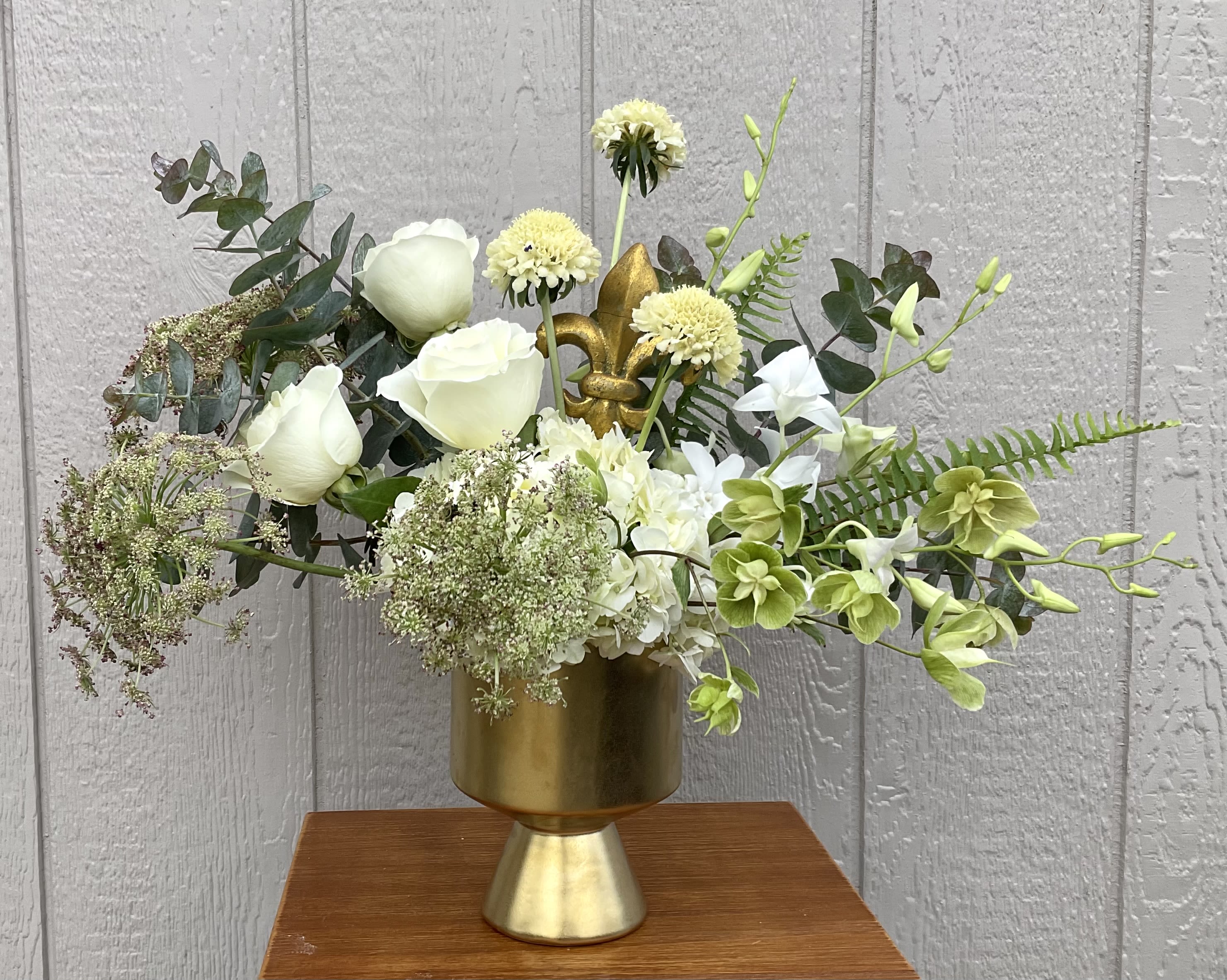 Parish Pearl - Soft white roses and pale green hellebores mingle with airy white hydrangea and delicate white orchids to create a calm, elegant flower arrangement perfect for a sympathy tribute, wedding décor, or a thoughtful housewarming gift. The Parish Pearl arrangement reads as a refined everyday bouquet and as an understated centerpiece for moments that call for quiet beauty-the person you're thinking of will feel soothed and seen by its soft palette and garden-like silhouette.  Arranged asymmetrically in a metallic gold vase, this design pairs branching stems and fine-textured filler for a loose, airy profile that feels modern yet natural. Colors are anchored in whites and fresh greens with mixed-color accents from the assorted blooms; the gold container and Fleur de Lis pik adds a subtle decorative pop without overpowering the flowers. Sized to display on mantels, dining tables, or bedside stands, it arrives ready to place and enjoy. For best longevity, keep water fresh and out of direct sunlight to help the roses, hydrangea, chrysanthemums, and orchids bloom their fullest.