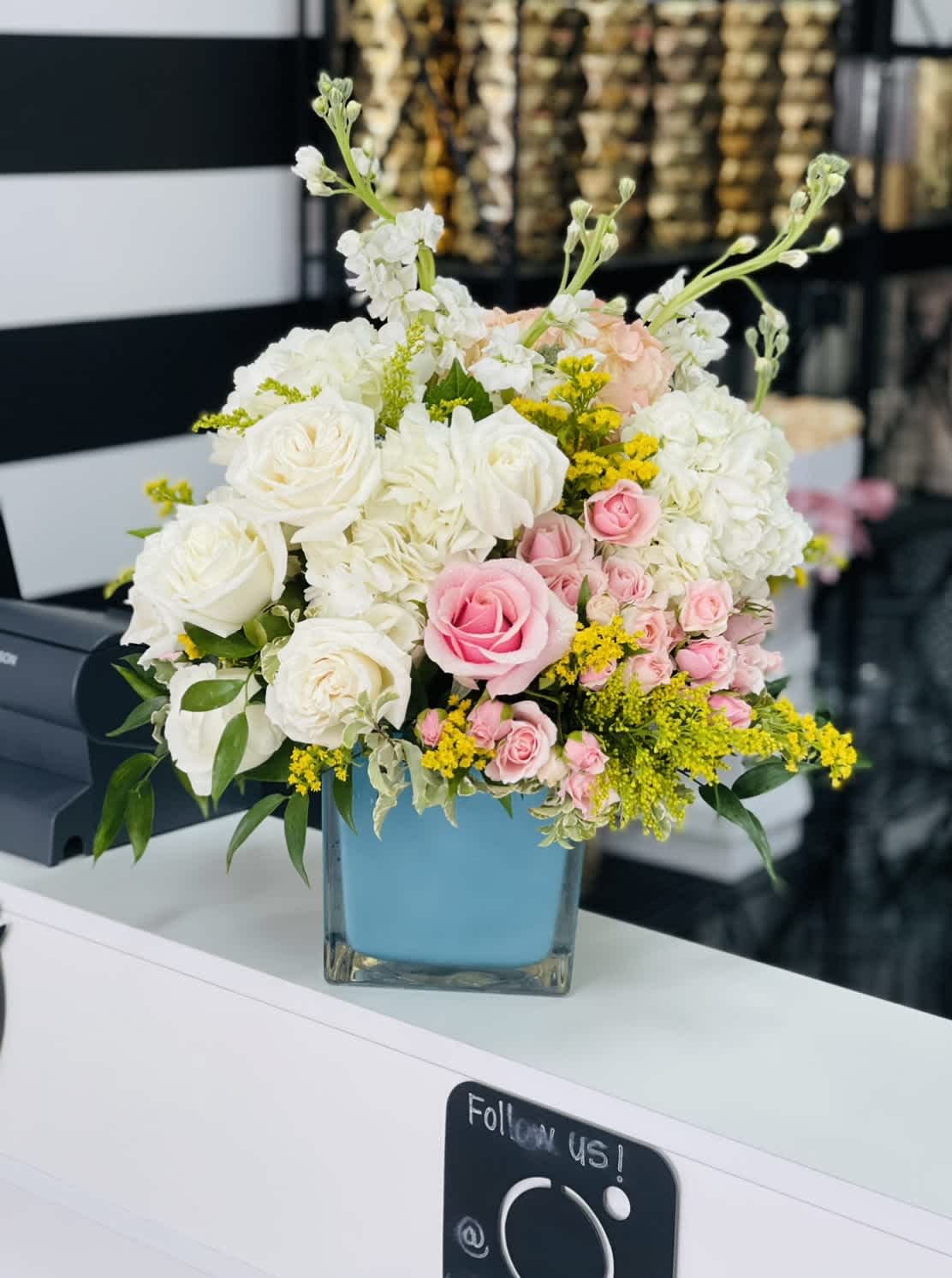  AQUA DREAM BOUQUET BY ATLANTA'S FINEST FLOWERS  - A soft cluster of blush-pink roses and creamy white hydrangea-like blooms framed by bright yellow asters makes a refreshing statement for a birthday bouquet or a spring and summer surprise. This flower arrangement-ideal for Mother's Day or to celebrate a special woman on Women's Day or Best Friends Day-lands as both romantic and cheerfully modern, the kind of gift that will make her feel truly seen.  The arrangement pairs white roses, light pink spray roses, white stock, and white hydrangea-style flowers with yellow aster accents and touches of green for texture. All stems are arranged in a polished 5" x 5" aqua cube (square glass) vase that echoes the cool blue-green tones and gives a contemporary finish. The finished size is approximately 10" x 10", a generous, rounded silhouette that fills a table without overwhelming it.  Perfect for birthdays, Boss's Day, Admin Professionals' Week, or as a Spring or Summer centerpiece, this bouquet arrives ready to display-simple water care will keep the blossoms fresh longer: change the water every couple of days and trim stems as needed. It's a graceful, uplifting choice when you want her to feel celebrated.