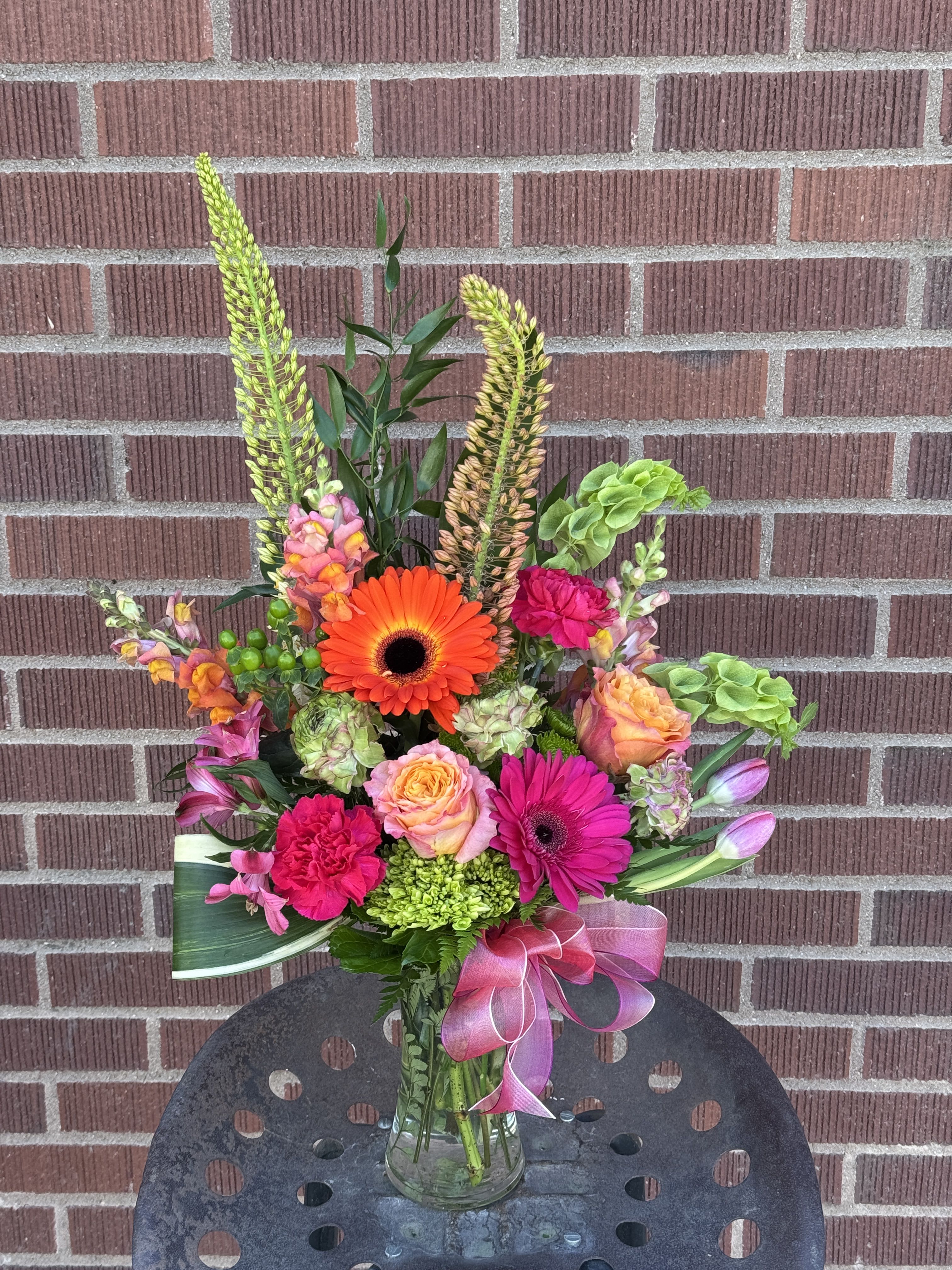 WEEKLY DROP - LIMITED EDITION! - Sunset Fiesta - Bring the warmth and energy of a radiant summer sunset indoors. This show-stopping design is a high-impact explosion of vivid coral, electric pink, and striking citrus tones. Foxtail lilies reach upward to create an elegant, architectural silhouette. Bold gerbera daisies instantly command attention and radiate joy. Sunset roses &amp; snapdragons add layers of rich, warm tones. Bright green hydrangeas &amp; bells of Ireland wrap up this high-energy piece.