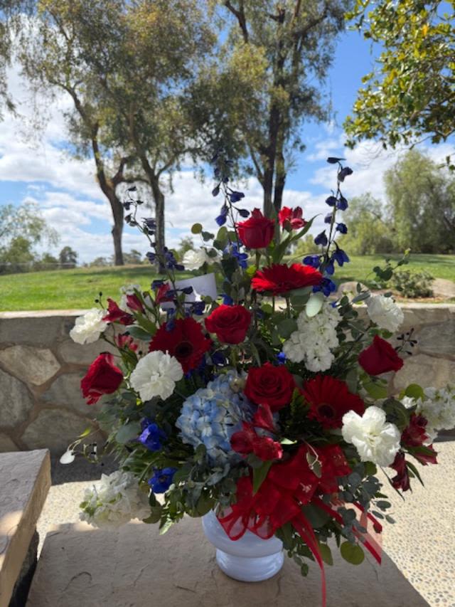 Patriotic Tribute  - This piece is designed tall and one sided for the service. It's designed in a white plastic urn style container that follows the national cemetery protocol. Flowers may include Delphinium, roses, carnations, hydrangea, lilies, alstroemeria, and/or stock depending on availability. It will be beautiful, in red white and blue.