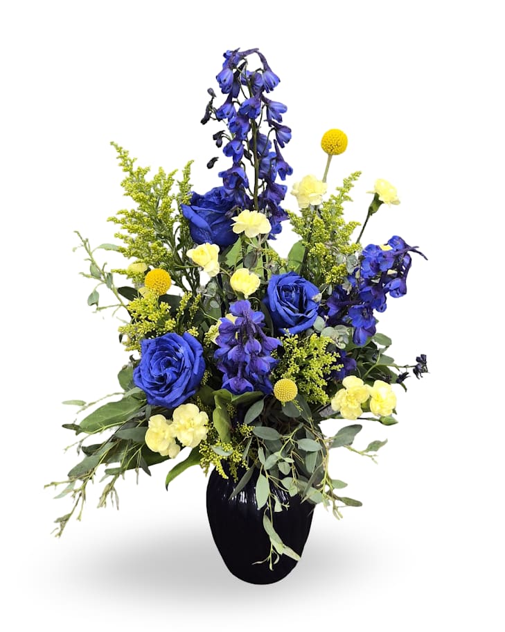 Blue &amp; Gold - Vivid cobalt-blue and sunny yellow blooms rise from a royal blue vase, the blue-and-gold color scheme creating a bold, celebratory statement perfect for graduation or a summer welcome for a new baby. Airy green foliage fans around the arrangement, giving it a layered, full silhouette that reads cheerful yet slightly formal-ideal for congratulations or a bright thank-you gift.  Designed to feel both festive and refined, this vase arrangement fits tabletops and reception desks for a bright touch.