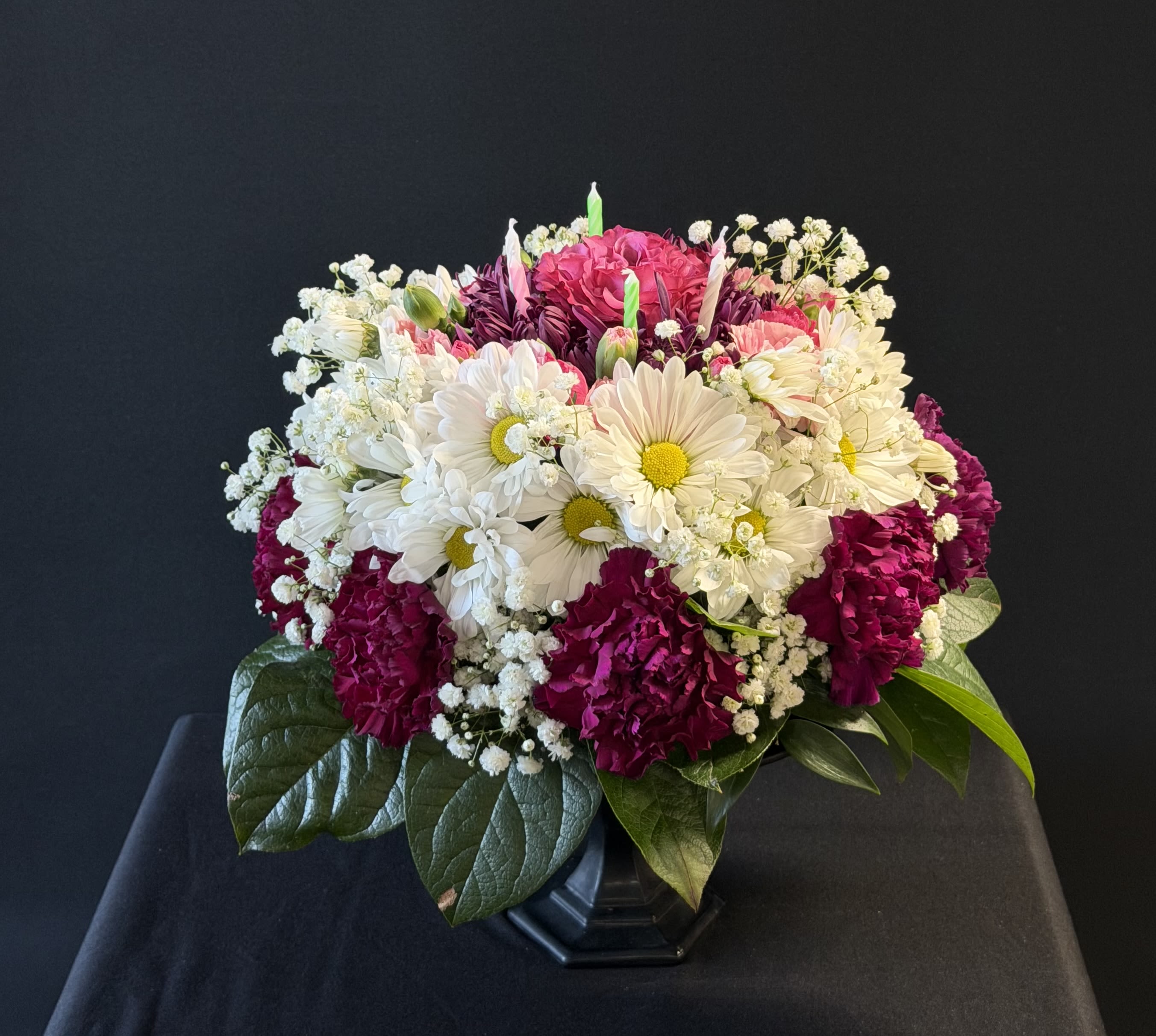 Floral Birthday Cake - A lively, full-bodied arrangement that reads like a floral celebration-ideal as a birthday bouquet to make someone feel especially seen. Bright white daisy-like blooms create a fresh, joyful foundation while saturated magenta carnations and pink and purple accents add bursts of cheer; the overall effect is playful, feminine, and unmistakably festive for a birthday or thank-you moment.  Soft clusters of baby's breath lend an airy, romantic texture around the edges, and glossy greenery lifts the color palette with cool green tones. Flowers are gathered in a sleek black vase that anchors the composition and lets the pinks, lavenders, and whites pop-perfect for gifting or to brighten a bedside table or kitchen island.  This arrangement suits recipients who love bright, happy color and classic garden blooms; it's the kind of gift that makes her feel celebrated and remembered. Vase-arranged for easy display-refresh water every few days and remove any wilting stems to extend enjoyment.