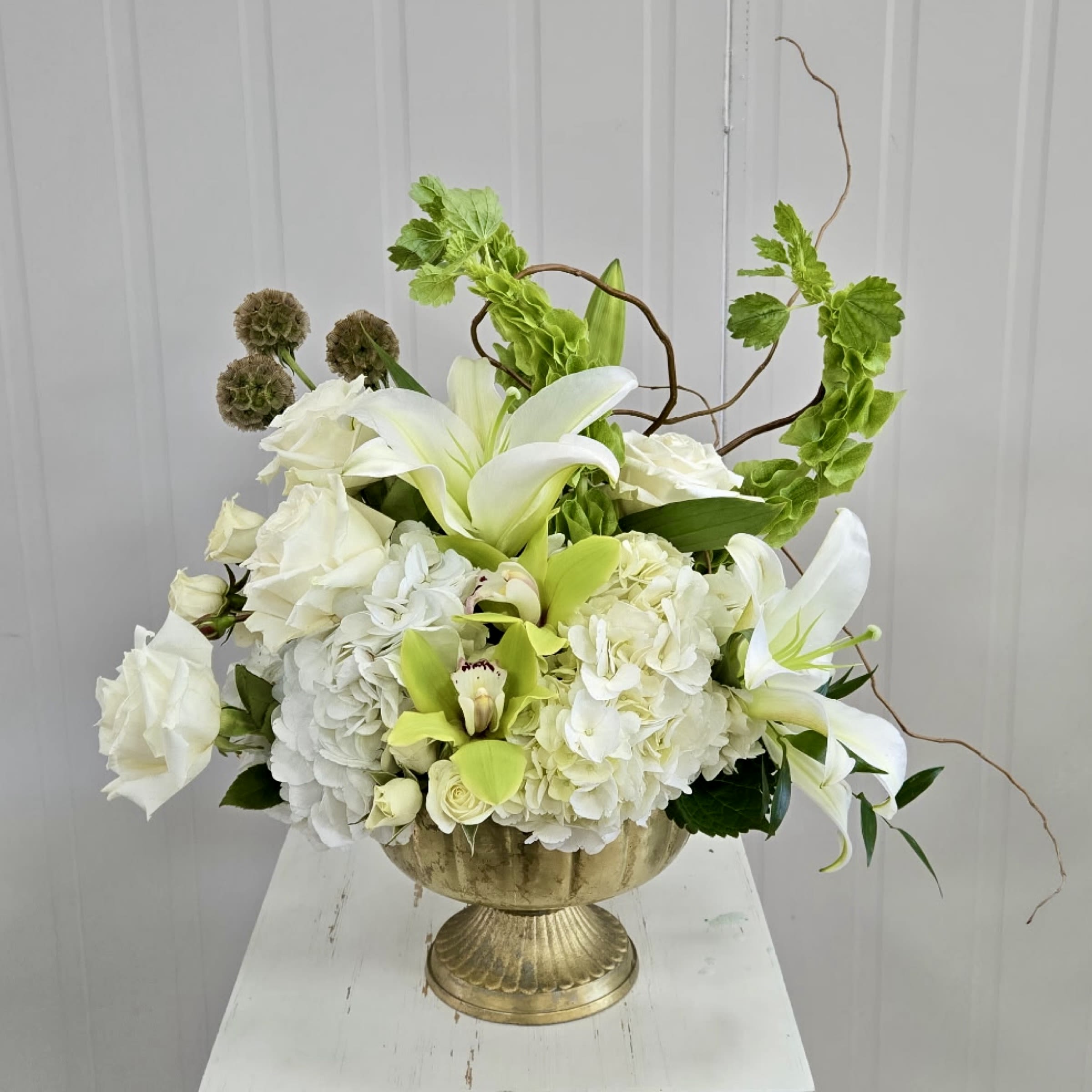 Lavish Luxury - This Gold Pedestal Dish Arrangement Includes White Roses, White Spray Roses, White Lily, Bells of Ireland, Scabiosa Pods, White Hydrangea and Green Cymbidium Blooms. Or as Similar as Possible. 