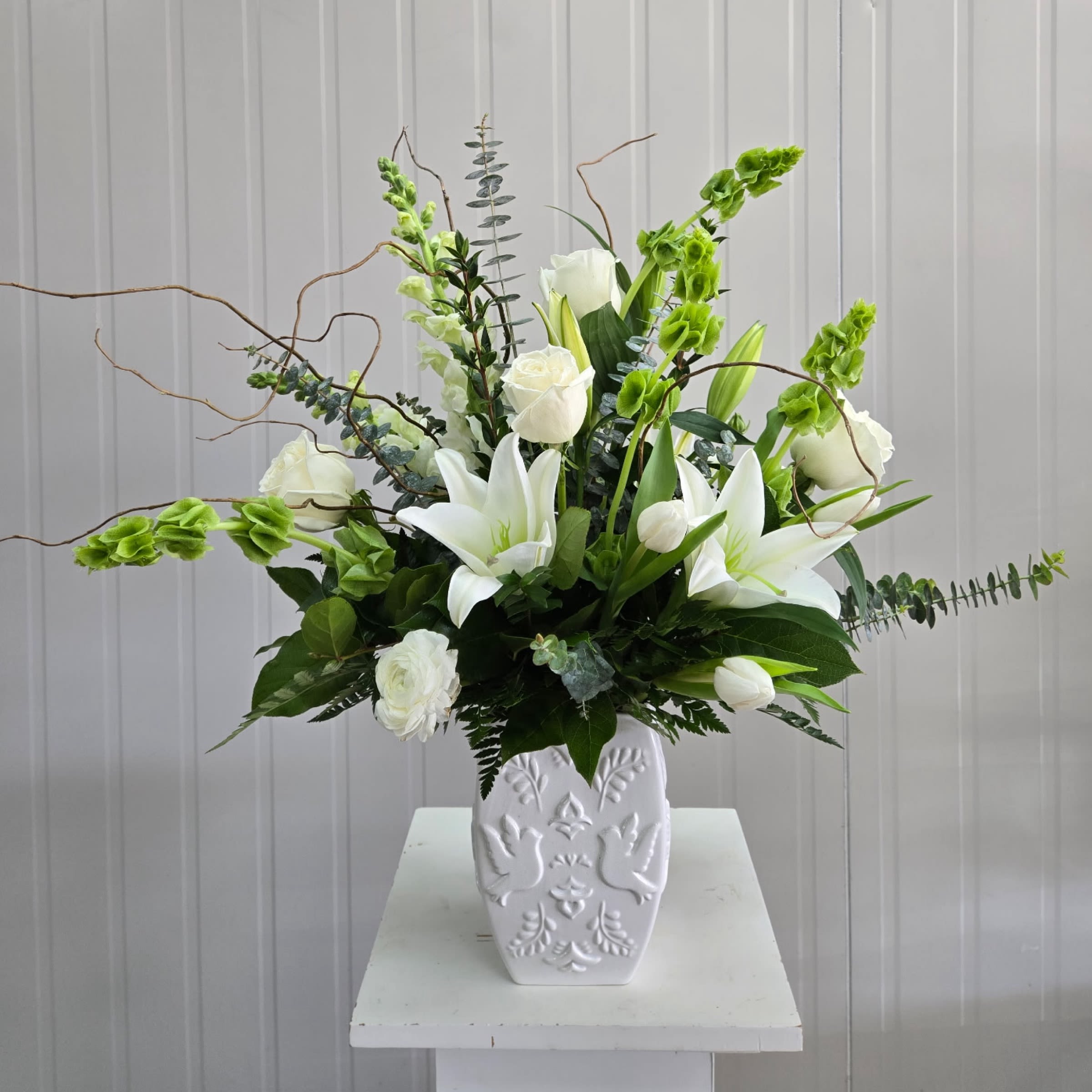 Wing of Comfort - This White Ceramic Vase Arrangement Includes White Lily, White Snapdragon, Bells of Ireland, White Rose, White Tulips or Other Seasonal Bloom with Curly Willow Accent Branches. Or as Similar as Possible. 
