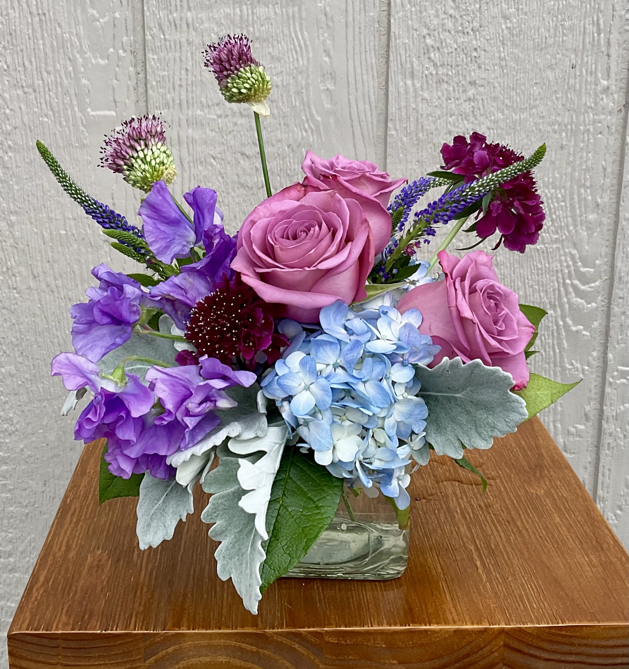 Twilight Garden - Lavender roses set against a rounded cluster of pale blue hydrangea give this flower arrangement a dreamy, garden-fresh look - perfect as a birthday bouquet, a thank-you gift, or a spring celebration arrangement. The mix of lavender, purple, blue, and white creates a cheerful yet gently romantic mood that will make the person you're thinking of feel noticed and celebrated.  This arrangement pairs lavender roses with pale blue hydrangea, several purple accent blooms, tall spiky stems for vertical interest, and silvery foliage for texture. Everything is arranged in a clear square glass vase that keeps the composition compact and tidy while showing off the stems. The layered colors and varied textures give it a lively, fresh scent profile and an elegant, hand-tied appearance.  Ideal for brightening a desk, bedside table, or entryway, Twilight Garden feels like a thoughtful everyday luxury that suits many occasions. Order for same-day delivery when available; keep the vase topped with fresh water and out of direct sunlight to extend the life of the blooms.