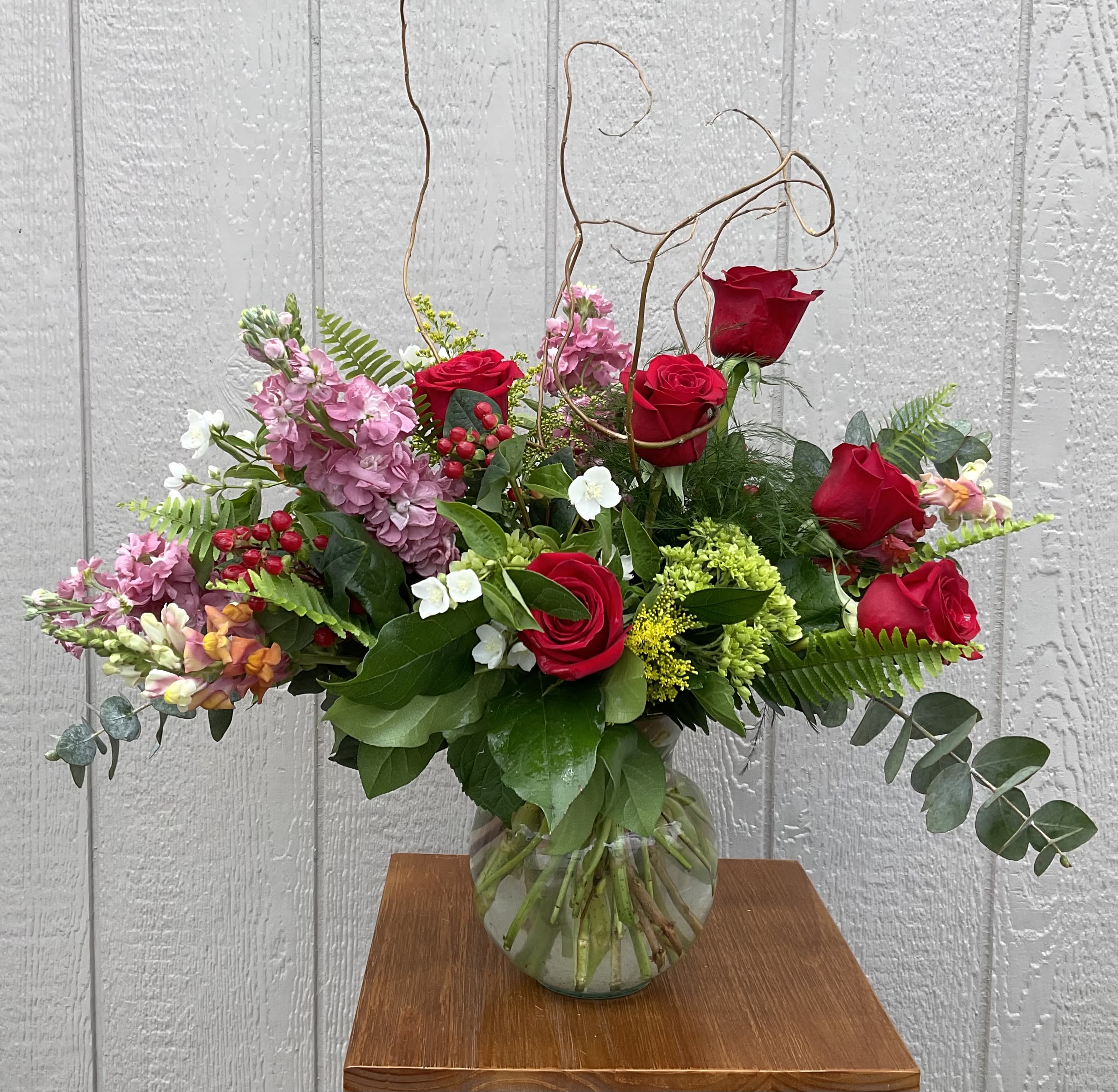 Saffron Sunset - Bright red roses and blushing pink stock create an immediately romantic, celebratory impression in this flower arrangement-perfect as a birthday bouquet, anniversary flowers, Valentine's Day surprise, or a heartfelt thank-you. The name suggests warmth, but the design reads fresh and garden-like: the recipient will feel seen and cherished the moment these rich reds and soft pinks arrive.  Clusters of red roses and pink stock are accented with small white blooms and abundant greenery gathered in a clear glass vase. The composition is broad and airy, with looping curly willow rising above the blooms and long fern- and eucalyptus-like foliage stretching outward for an elegant, asymmetrical silhouette. The mix of textures-velvety rose petals, feathery foliage, and delicate stock-gives the arrangement substance without feeling heavy.  This substantial everyday arrangement suits many celebratory moments and makes a striking centerpiece on delivery day; it arrives ready to display in the included clear glass vase. For best enjoyment, keep flowers in a cool spot away from direct sun, refresh the water every couple days, and trim stems slightly when you change the water to help the bouquet last.
