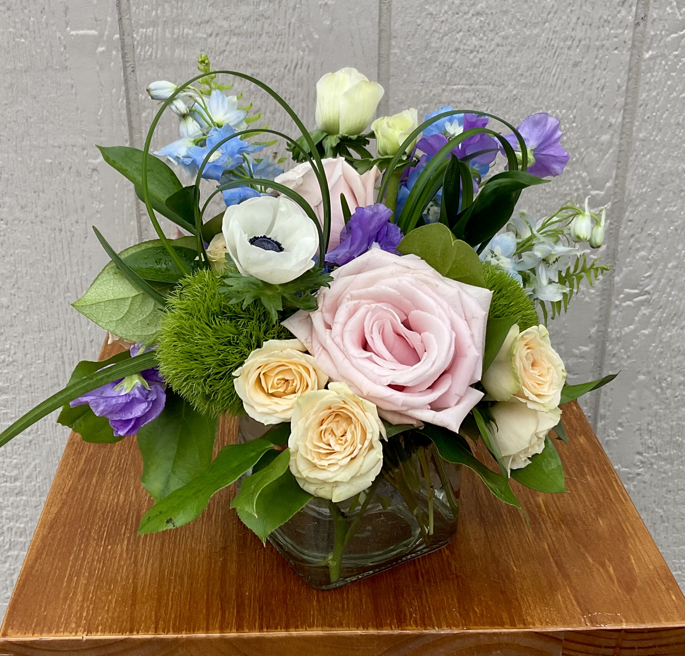 Serene - Large blush-pink roses takes center stage in this Serene flower arrangement, setting a soft, romantic tone perfect for a birthday bouquet, a cheerful thank-you, or an everyday gift to make someone feel truly noticed. The combination of pink, white, purple, and blue gives the arrangement a fresh, pastel palette that reads as calming and celebratory-an ideal choice when you want the recipient to feel comforted and uplifted.  This full, rounded design pairs blush roses with cream spray roses, white anemones, airy delphinium and lisianthus accents, plus other delicate blooms for layered texture. Looping grass-like stems and broad green leaves add movement and a natural, garden-fresh feel, while the clear glass vase keeps the presentation simple and elegant so the mixed pastels can shine. Sized to sit beautifully on a dining table or desk, Serene arrives ready to brighten a room and the day of the person you're thinking of.