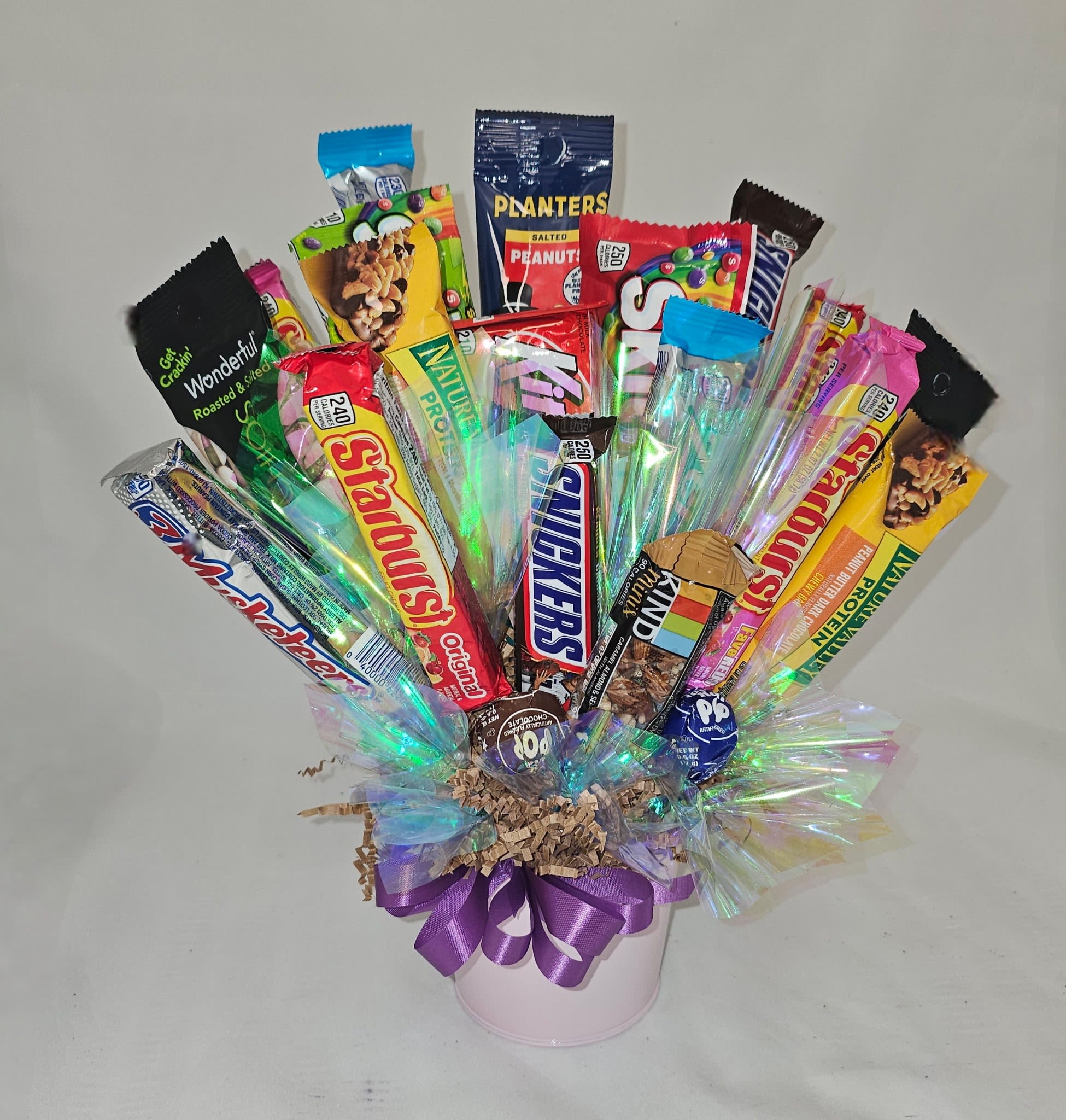 Candy Basket - A playful candy bouquet that feels like a snack-lover's party in a container - perfect as a birthday gift, casual thank-you, or cheerful pick-me-up. This candy basket-style arrangement presents assorted candy and snack bars fanned into a compact bouquet that reads bright and celebratory, so the person you're thinking of will immediately feel indulged and noticed.  Wrapped in clear iridescent cellophane with a bow, the mix of purple, pink, black, and mixed-color packaging creates glossy, eye-catching texture. Shredded paper filler keeps the bars neatly displayed in the container so the presentation stays tidy from unboxing through snacking. No floral foam or stems - just edible treats artfully arranged for instant enjoyment.  Ideal for coworkers, friends, teens, or anyone with a sweet tooth, this cheerful snack basket sends a lighthearted message of celebration or thanks. Order with confidence for same-week gifting; no special care needed beyond keeping the arrangement dry and away from heat to protect the candy.
