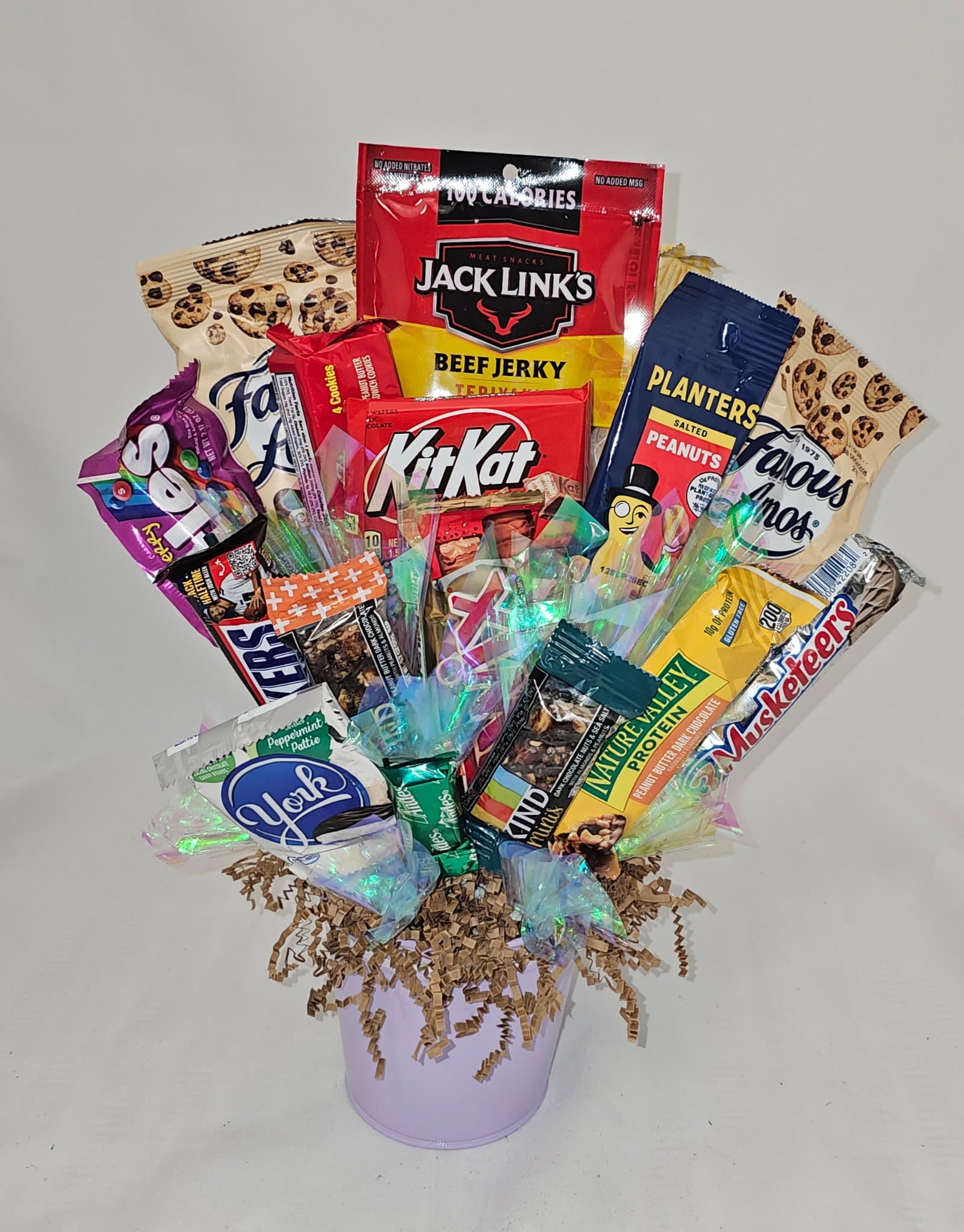 Snack Basket - A playful snack gift basket that reads like a bouquet-perfect for birthday celebrations, teacher appreciation, or a fun "thinking of you" surprise. This snack arrangement is presented in a container and styled like a casual bouquet so the recipient feels delight and lighthearted surprise the moment they see it.  Assorted packaged snacks and candy stand upright amid crinkled paper filler, their red, blue, yellow and tan wrappers layered to create a bright, busy composition that fans outward like stems in a bouquet. The container and crinkled paper add texture and volume while keeping everything neatly contained and ready to gift; the arrangement leans informal and whimsical rather than floral, making it ideal for anyone who prefers treats over traditional flowers.  Send this to celebrate a milestone, say thanks, or bring a smile to a friend or coworker - it's the sort of gift that feels personal and immediate. Ready to hand over as-is, no assembly required; simply unwrap and enjoy the snack variety inside.
