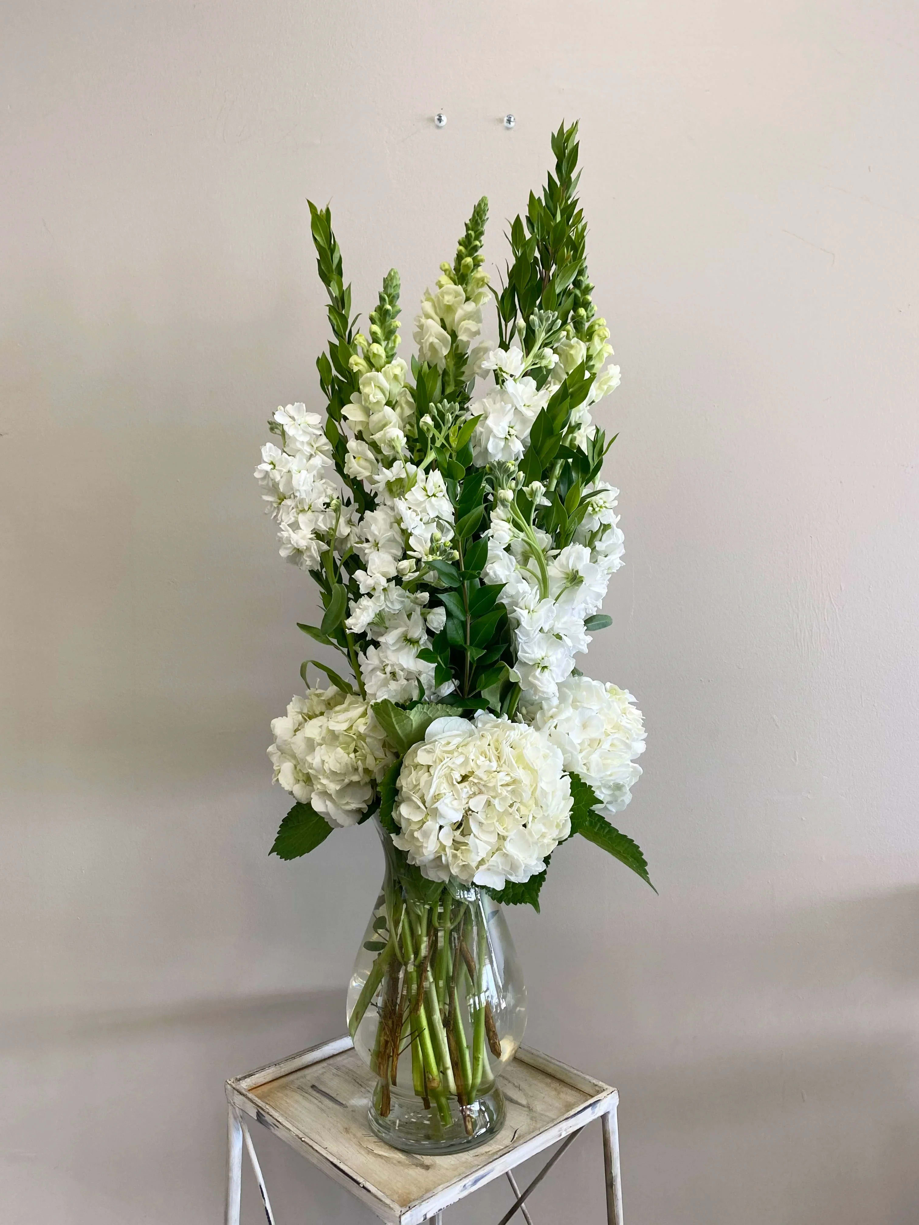 Tall white hydrangeas and snapdragons arranged in a clear glass vase on a small wooden stand