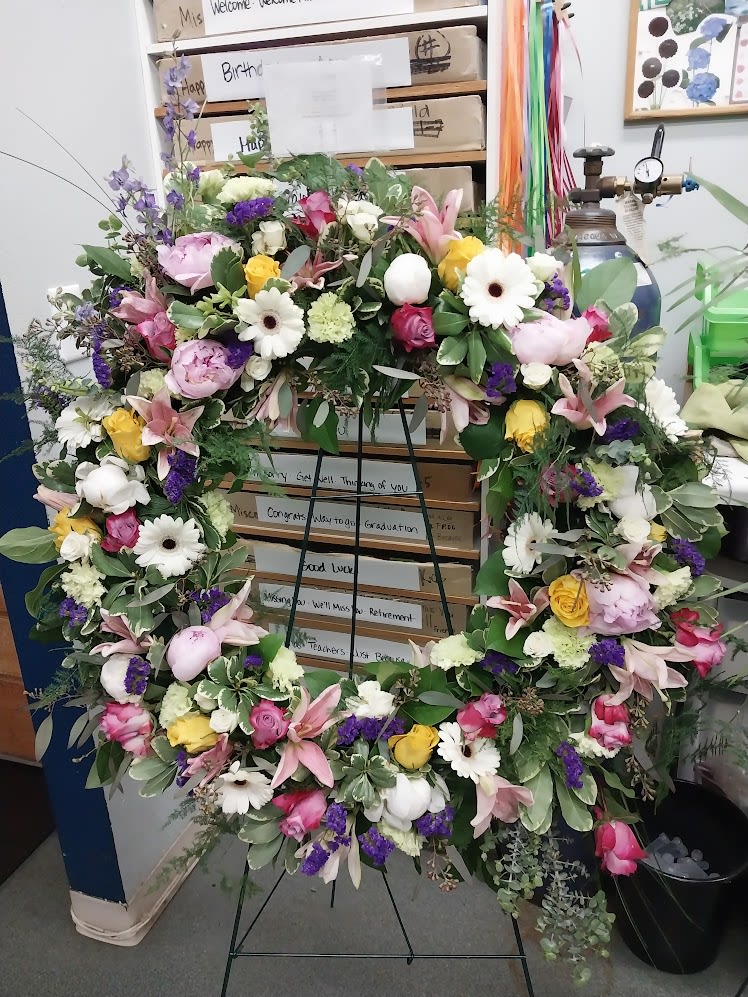 Love's Lasting Circle - A lush garden-inspired wreath featuring roses, lilies, and seasonal blooms in soft pink, lavender, cream, and yellow tones.