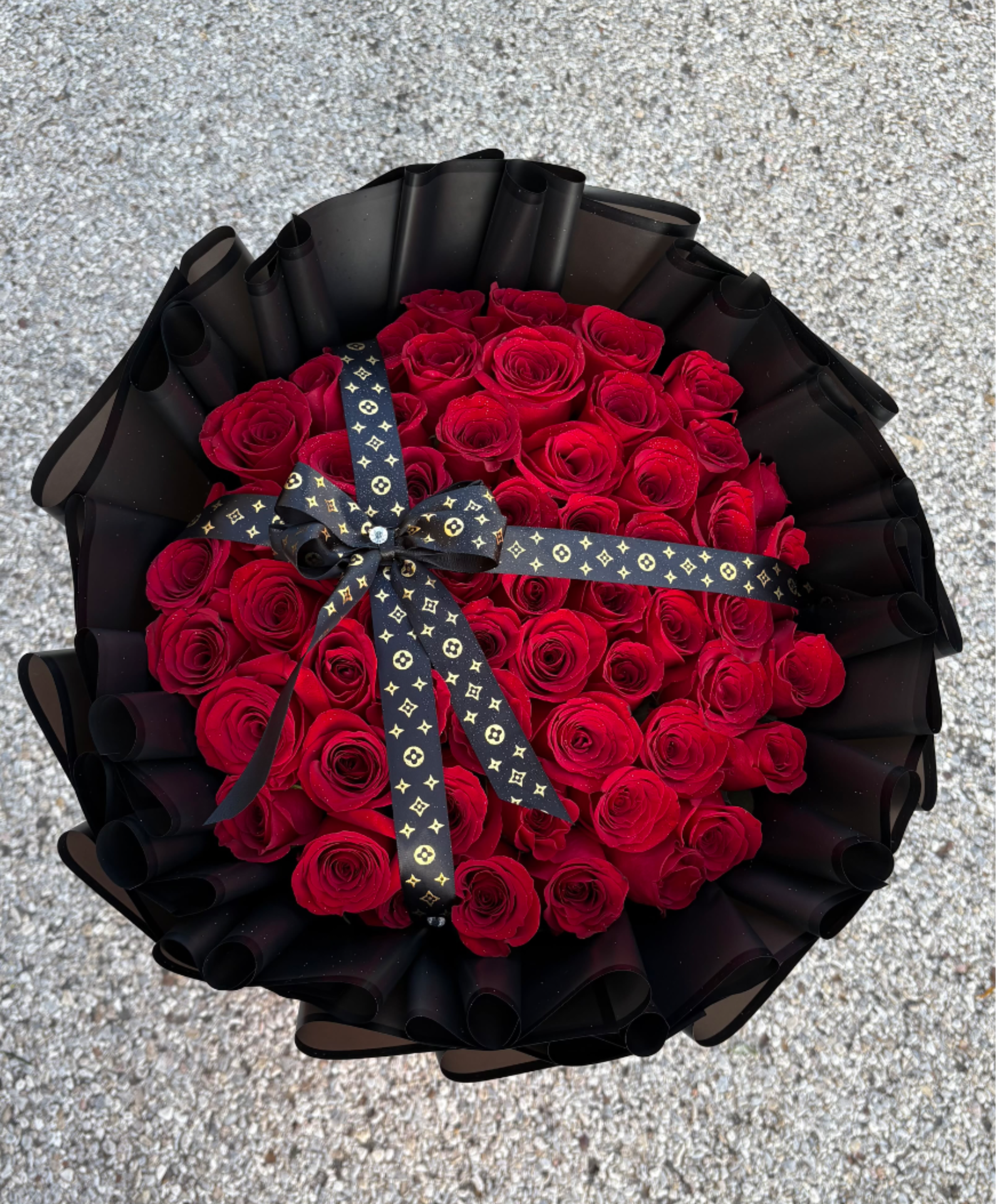 LUX RED - A dense dome of 50 red roses arranged in a classic rounded bouquet makes a powerful romantic statement-ideal as an anniversary bouquet or a Valentine's Day gift for the person you want to sweep off their feet. The vivid scarlet blooms are tightly packed to create a lush, full head of roses that reads as both luxurious and intensely romantic when paired with the dramatic black presentation.  Wrapped in layered black paper with curled edges and finished with a black ribbon printed with a gold monogram-style pattern, the contrast between the deep red petals and the dark wrap gives the bouquet a bold, formal look that feels upscale and intentional. The arrangement arrives ready to gift; stems are gathered and secured beneath the swirl wrapping to hold the domed shape-she'll feel truly celebrated the moment it's handed to her.