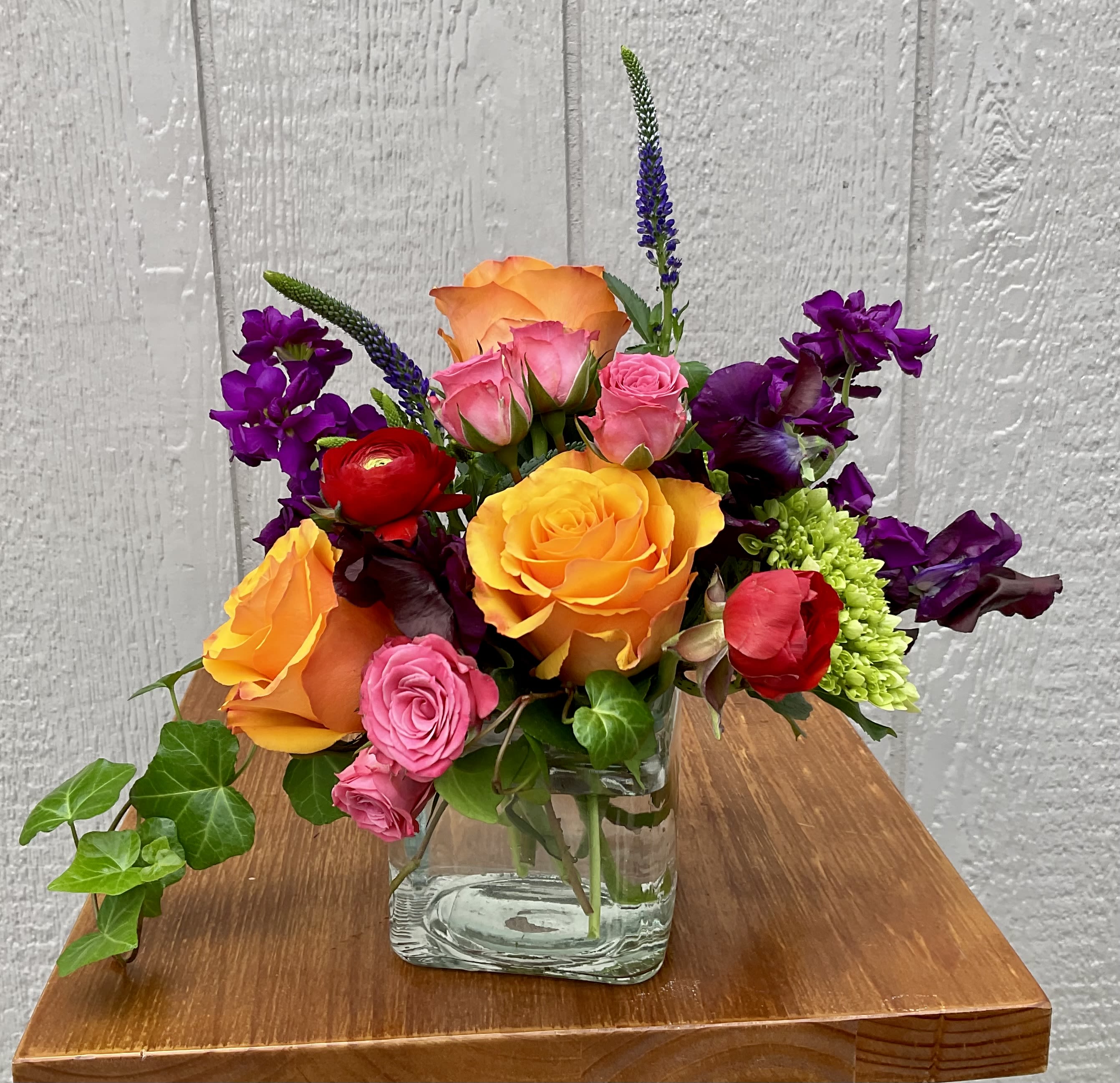 Revel - A vivid, celebratory bloom mix designed to brighten any special moment - Revel is a mixed-color flower arrangement perfect for birthdays, congratulations, or simply to say "I'm thinking of you." Plush roses in bright orange, pink, red and yellow form a dense, joyful cluster that immediately reads as festive and heartfelt, while tall purple and green spiked flowers add vertical energy and contrast for an eye-catching silhouette.  This compact arrangement sits in a clear square glass vase that keeps the design modern and neat, letting the stems show through for a fresh-cut garden feel. Alongside roses and stock you'll notice textural accents and a few trailing ivy leaves that soften the edges and add movement. The palette of saturated pinks, purples, oranges and warm yellows creates an exuberant, mixed-color statement that works equally well for celebratory tables, office desks, or a bedside surprise.  Revel is the kind of bouquet that makes the recipient feel genuinely seen and celebrated - bold, cheerful, and impossible to ignore. Its upright, compact shape fits small to medium spaces; refresh stems with clean water every couple of days and recut if you wish to extend vase life. Order for same-day or scheduled delivery to make their day memorable.