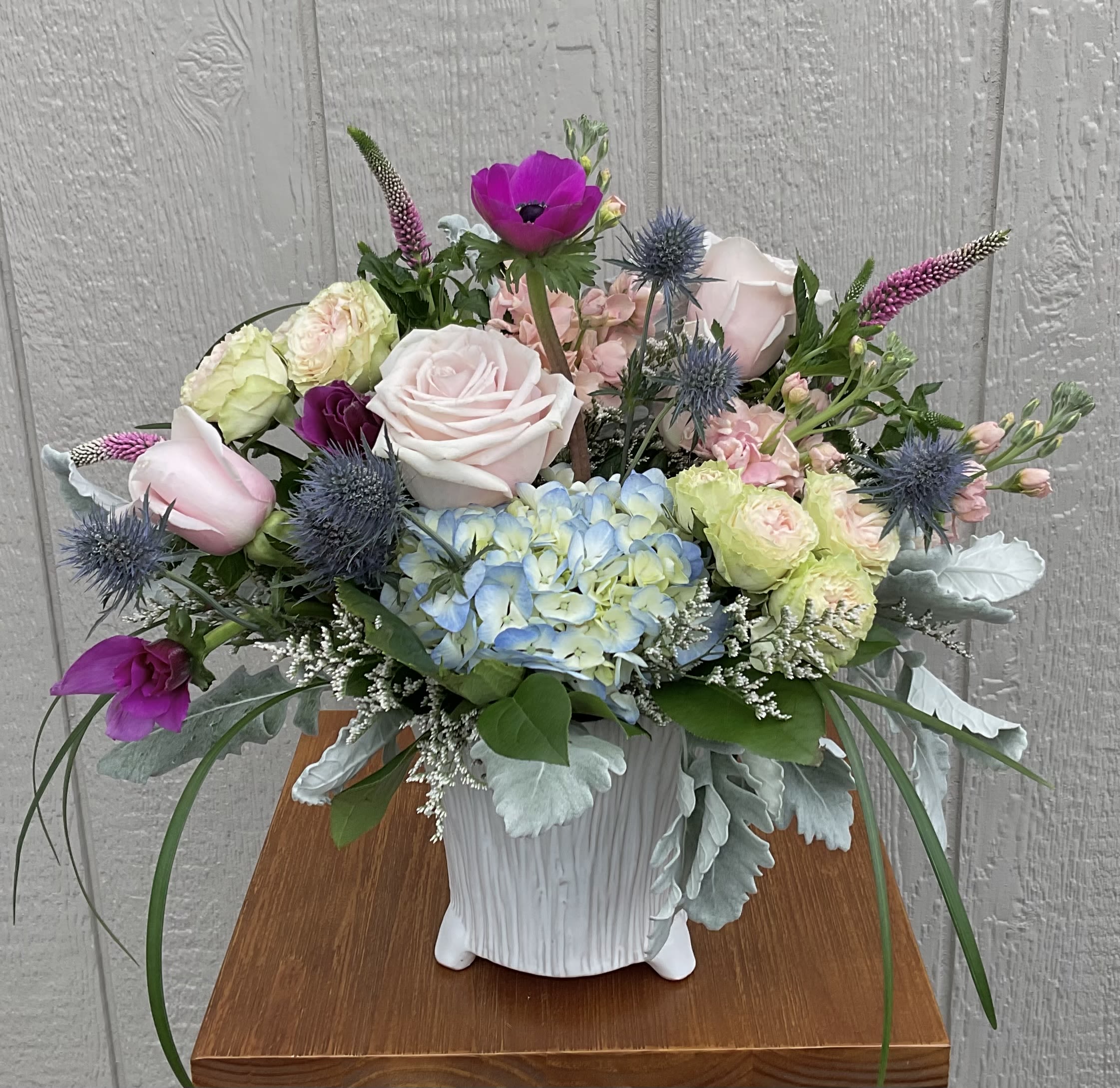 Prytania - Soft pink and cream roses mingle with a bold blue hydrangea cluster in this rounded flower arrangement-perfect as a spring birthday bouquet, a cheerful thank-you gift, or an everyday pick-me-up. The combination of anemones and vivid purple blooms with spiky blue thistle-like flowers gives the Prytania arrangement a lively, romantic presence that will make the person you're thinking of feel seen and celebrated.  This composition sits in a white textured ceramic vase and is finished with pale silver decorative foliage for added texture and modern contrast. Airy branching accents lift the bouquet, creating a full, garden-fresh silhouette that looks generous without feeling fussy. Soft pastels balanced by pops of blue and purple make it ideal for moms, friends, or anyone who appreciates elegant colors and a contemporary vase they can reuse.  Choose a size that fits your moment-a fuller arrangement fills a tabletop while a standard size brightens a desk or bedside. To keep blooms looking their best, refresh the vase water every few days and trim stems slightly when you change the water; these simple steps help ensure the roses, hydrangeas, tulips, and accents stay vibrant through the week.