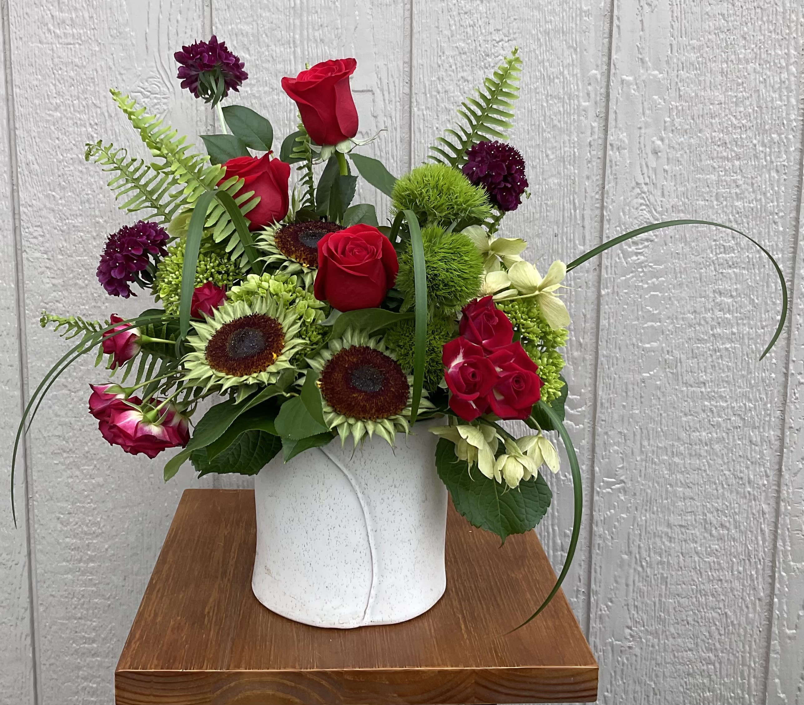 Scarlet Sage - Bright scarlet roses and small sunflower heads give this flower arrangement an immediate burst of color - a lively everyday bouquet perfect for birthdays, anniversaries, or to celebrate someone special. The mix of red, green, purple, and cream reads both joyful and slightly dramatic, the kind of gift that makes the person you're thinking of feel genuinely seen and celebrated.  The design pairs rich red roses with deep purple dianthus and pale cream accent blooms arranged in a smooth white ceramic vase. Long looping blades and airy fern fronds create an asymmetrical, garden-style silhouette that feels full and natural rather than overly formal. The compact white vase keeps the focus on the blooms while adding a clean, modern touch to tabletops or entryway consoles.  This arrangement suits anyone who loves bold color and textured, hand-tied styling - ideal for marking a birthday, to say congratulations, or simply to brighten someone's day. Vase-ready and arranged by hand, it arrives full and fresh; water daily and trim stems if needed to extend vase life.