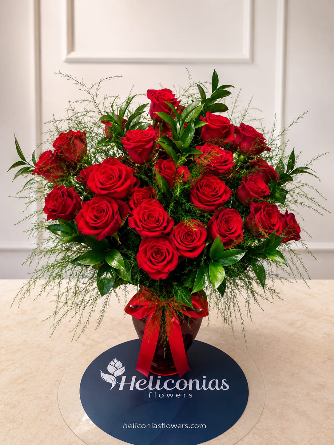 Eternal Romance red - A lush cluster of deep red roses creates an immediate, romantic impact-perfect as an anniversary bouquet or a heartfelt Valentine's arrangement to show the person you love how deeply you care. The rounded, upright design reads formal and timeless, making it equally appropriate for intimate celebrations or as a reverent sympathy display that communicates respect and warmth.  This arrangement combines rich red roses with fine, airy greenery and glossy foliage for contrast and texture. The flowers are gathered tightly in a dark glass vase finished with a wide red ribbon, presenting a polished, gift-ready look. Colors: red, green, and touches of white from the foliage highlights; hard goods: glass vase and ribbon.  Made to make someone feel truly seen and cherished, this is the kind of bouquet that says "I love you" with classic elegance or offers steady comfort in quiet moments. Order with vase delivery for a ready-to-display piece; to keep the roses fresh, change the water every other day and trim stems slightly when you refresh the vase.