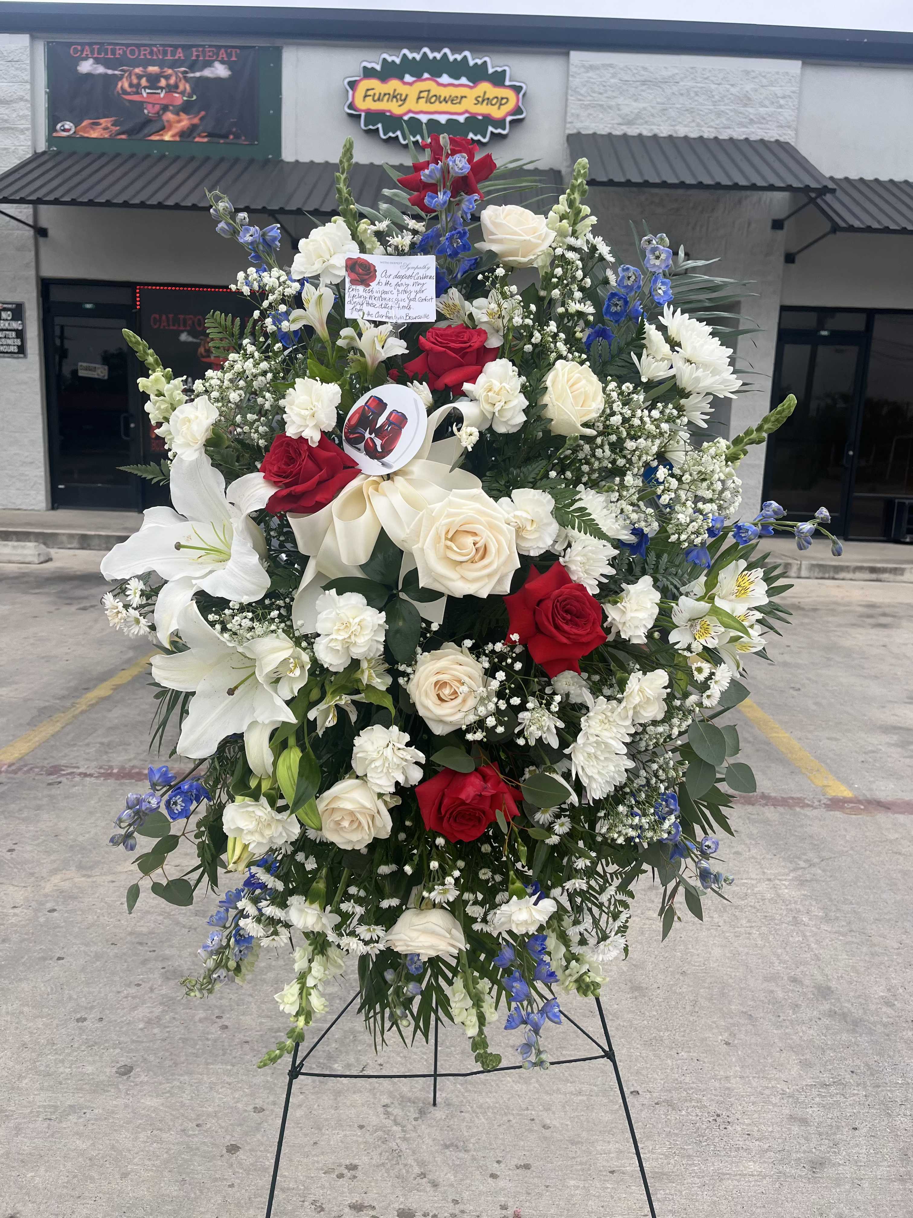 ode to you - A lush standing spray that reads like a heartfelt tribute, perfect as a sympathy arrangement to honor a loved one. This memorial spray combines red and white roses with white lilies to create a solemn, elegant focal point-the kind of floral tribute that helps the person you're thinking of feel remembered and respected.  Layered in a dense oval shape, the arrangement includes carnations, daisies, snapdragons and clusters of small filler blooms with blue accent flowers scattered through the mostly white palette for subtle contrast. Mounted on a black metal easel and finished with a decorative ribbon, the spray arrives with small inserted cards for a message or service detail. The overall dimensions are generous, designed to stand prominently at a ceremony or beside a grave site.  A meaningful way to express sympathy or pay tribute at a funeral or memorial service, this piece sends a clear message of love and remembrance. Care is simple-kept upright on its easel and away from direct heat-so it maintains its fresh, composed look through services and viewings.
