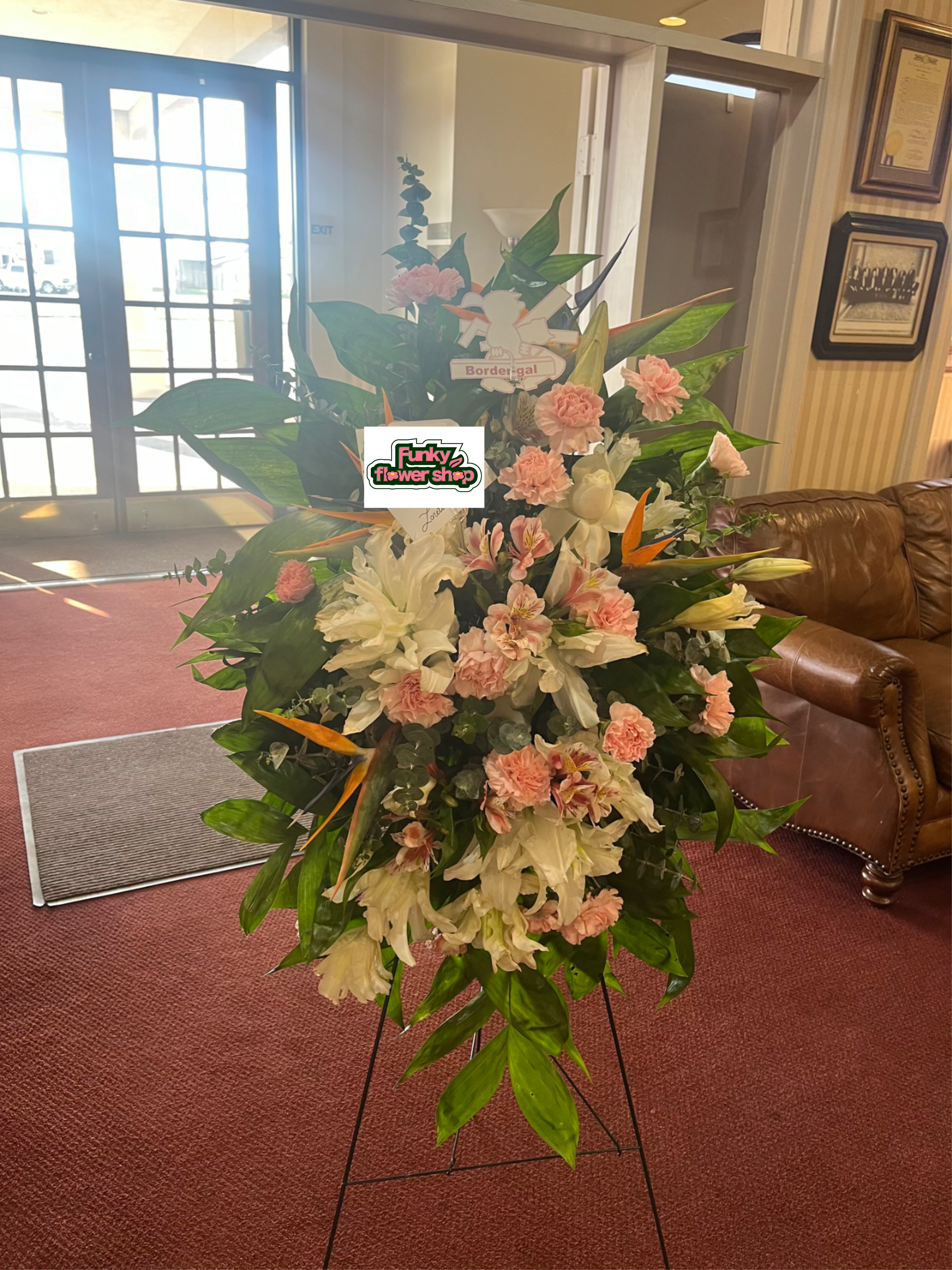 Graceful Horizon  - A lush, formal sympathy standing spray that speaks of comfort and reverence-this tall arrangement of white lilies, pink carnations, and vibrant orange tropical blooms is designed for funeral and memorial services. The metal easel stand and included sympathy card present the flowers with a ceremonial presence, making it a fitting tribute when you want the person you're thinking of to feel honored and remembered.  The design layers broad green foliage with clustered lilies and carnations through the center and lower half, while taller tropical accents, including striking bird-of-paradise, rise above for dramatic height and visual flow. Soft pinks and pure whites mingle with orange highlights and green leaves to create contrast and warmth; the composition is full and intentionally balanced to read well from a distance in a chapel or reception area.  Made to stand tall on its metal easel, this arrangement arrives ready for display at a service and includes a sympathy card for your personal message. Its scale and classic color mix convey respect and consolation-an understated yet powerful way to say you care.
