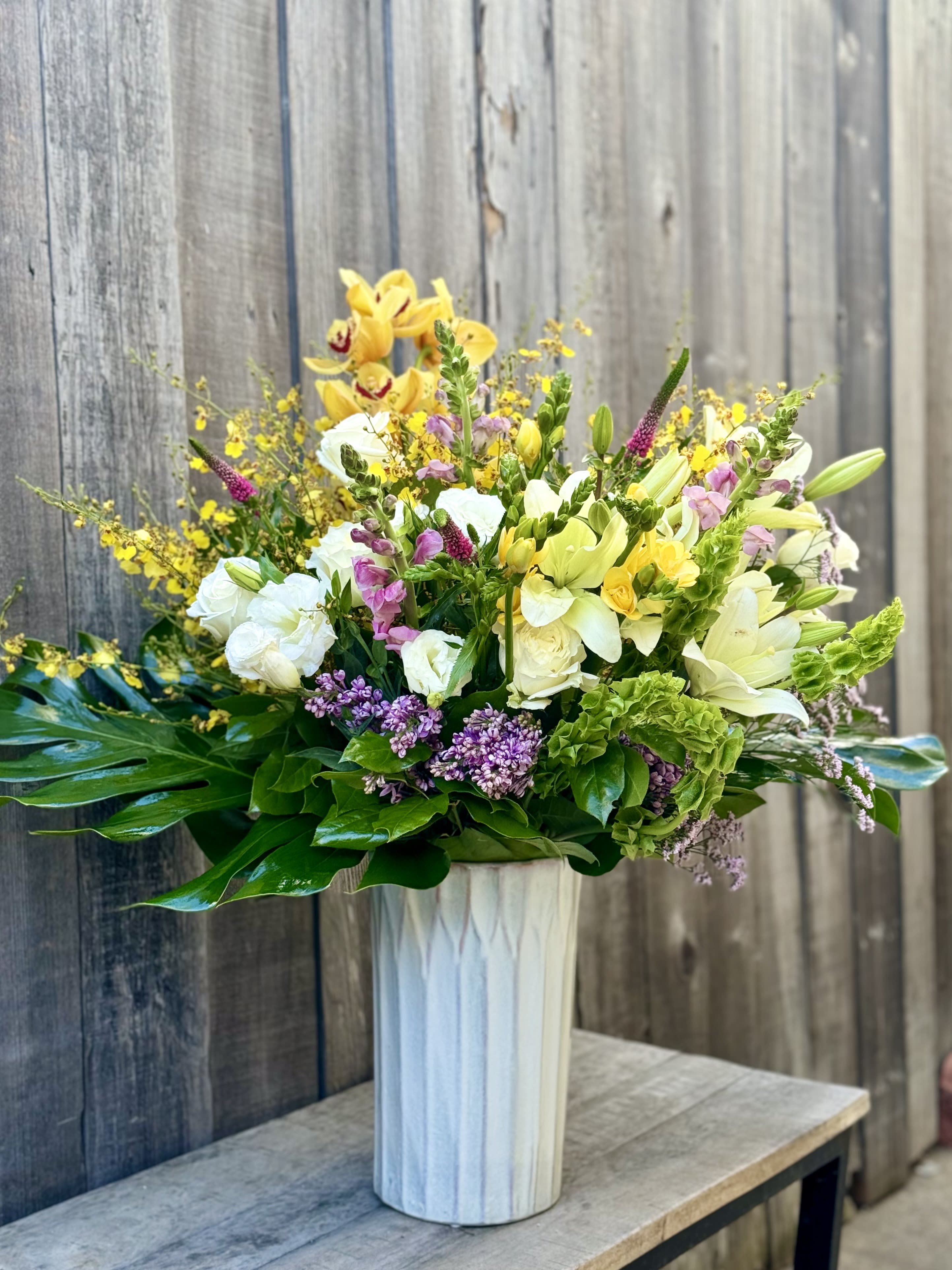 Mixed bouquet in a tall white vase with yellow, white, pink, and purple flowers.