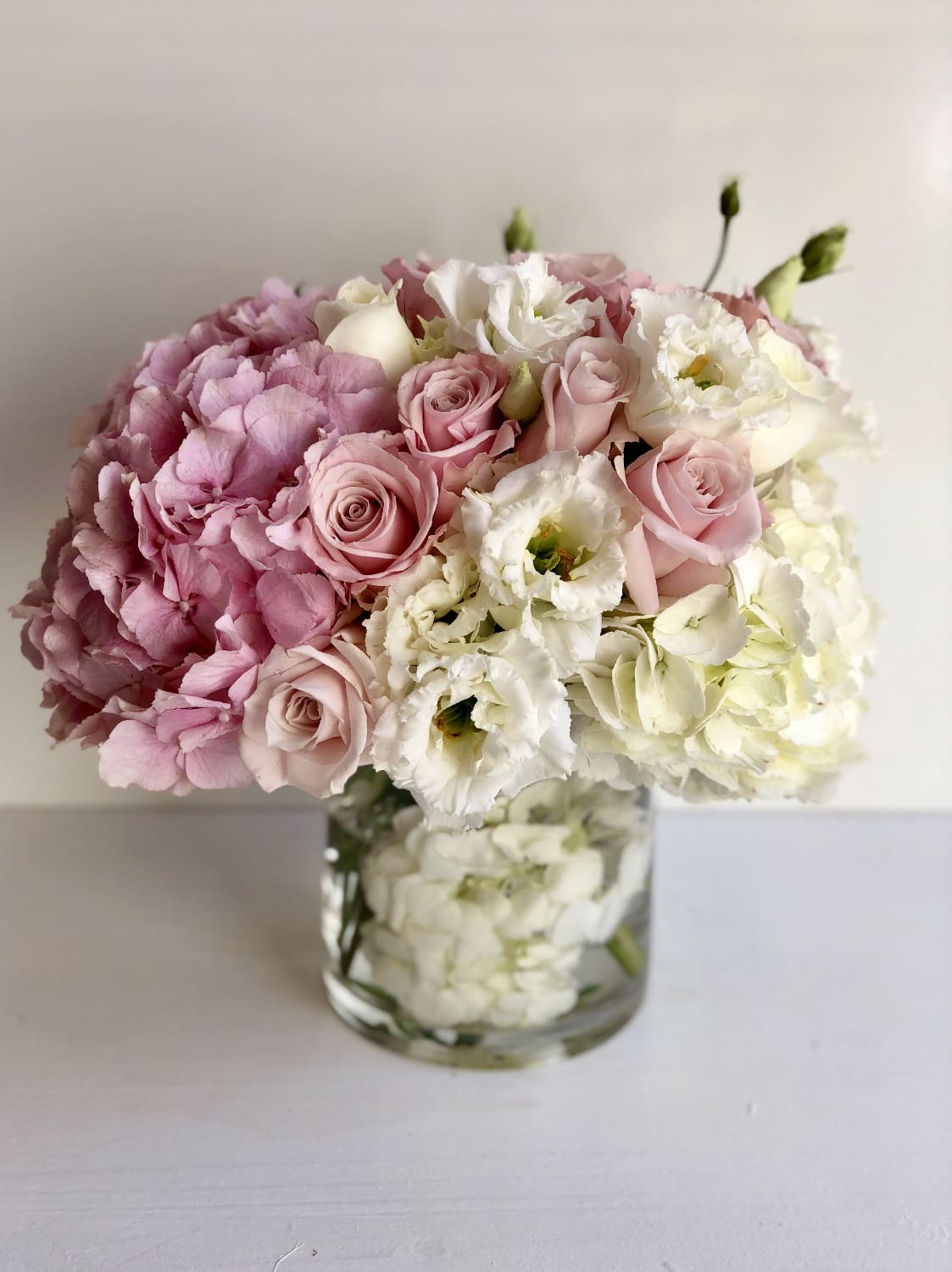 Low round arrangement of pink hydrangeas, pale pink roses, and white flowers in a clear glass vase.