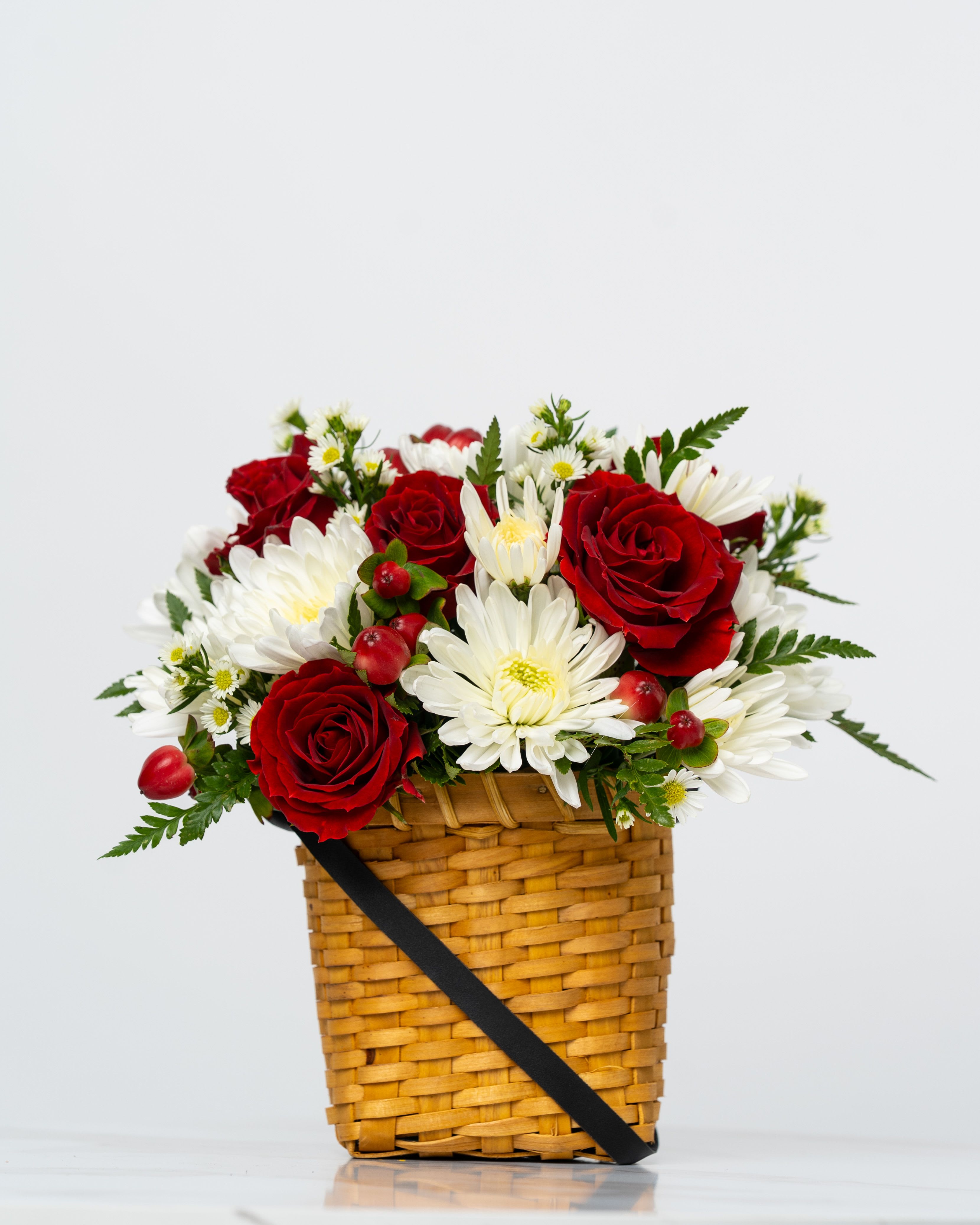 Red roses and white daisies in a woven basket with a black ribbon