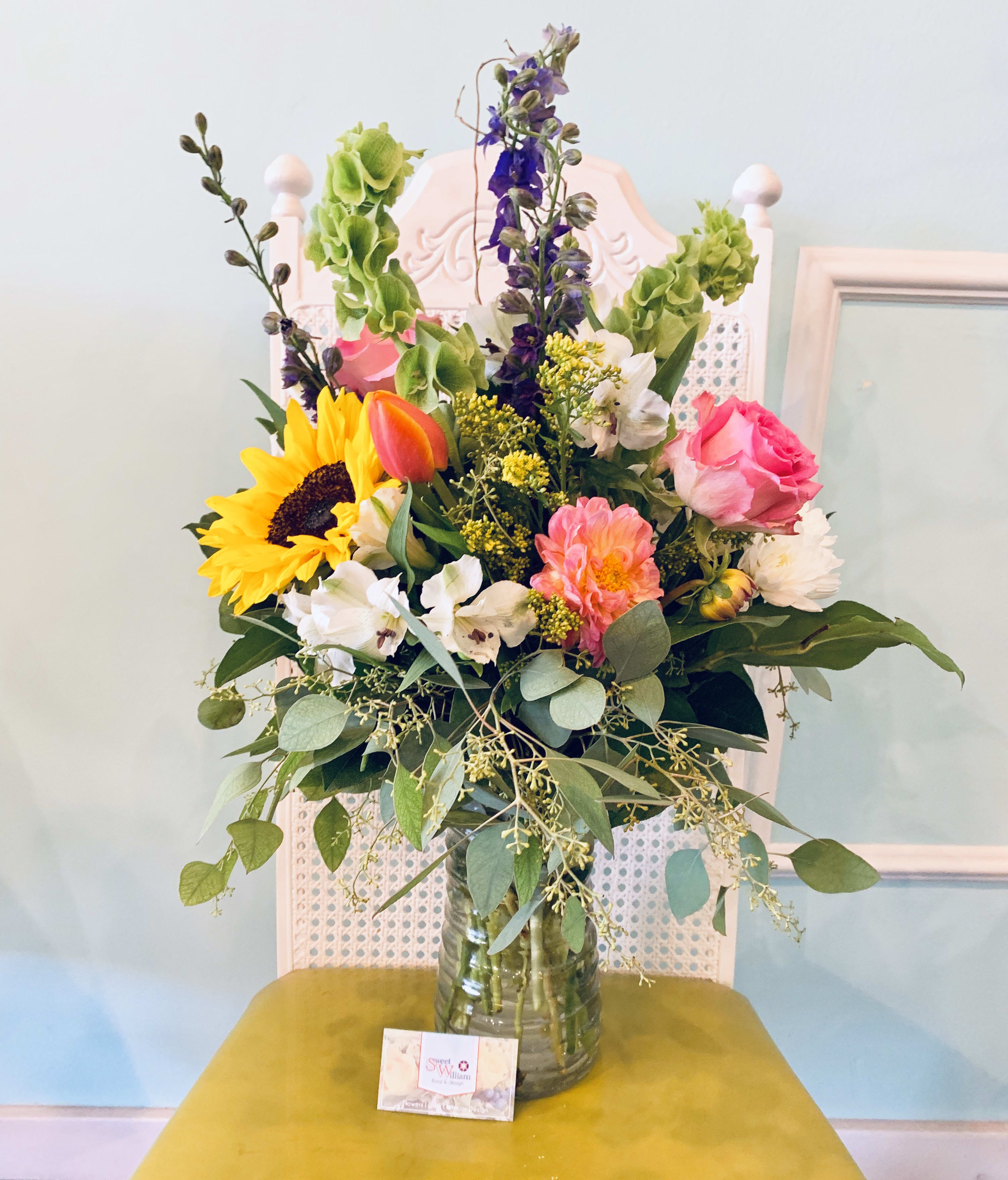 Here Comes the Sun - This arrangement full of bright and cheery colors will have you looking ahead to warmer days. Here Comes the Sun has sunflowers, bells of Ireland, blue delphinium, alstroemeria, and roses as well as seasonal flowers and greens that will have you walking along Abbey Road and humming this classic Beatles song.   Please Note: Standard Delivery for this product occurs in the afternoon, between 2:30pm and 7pm. If you need it by a certain time, there is an option to select a timed window for scheduled deliveries.