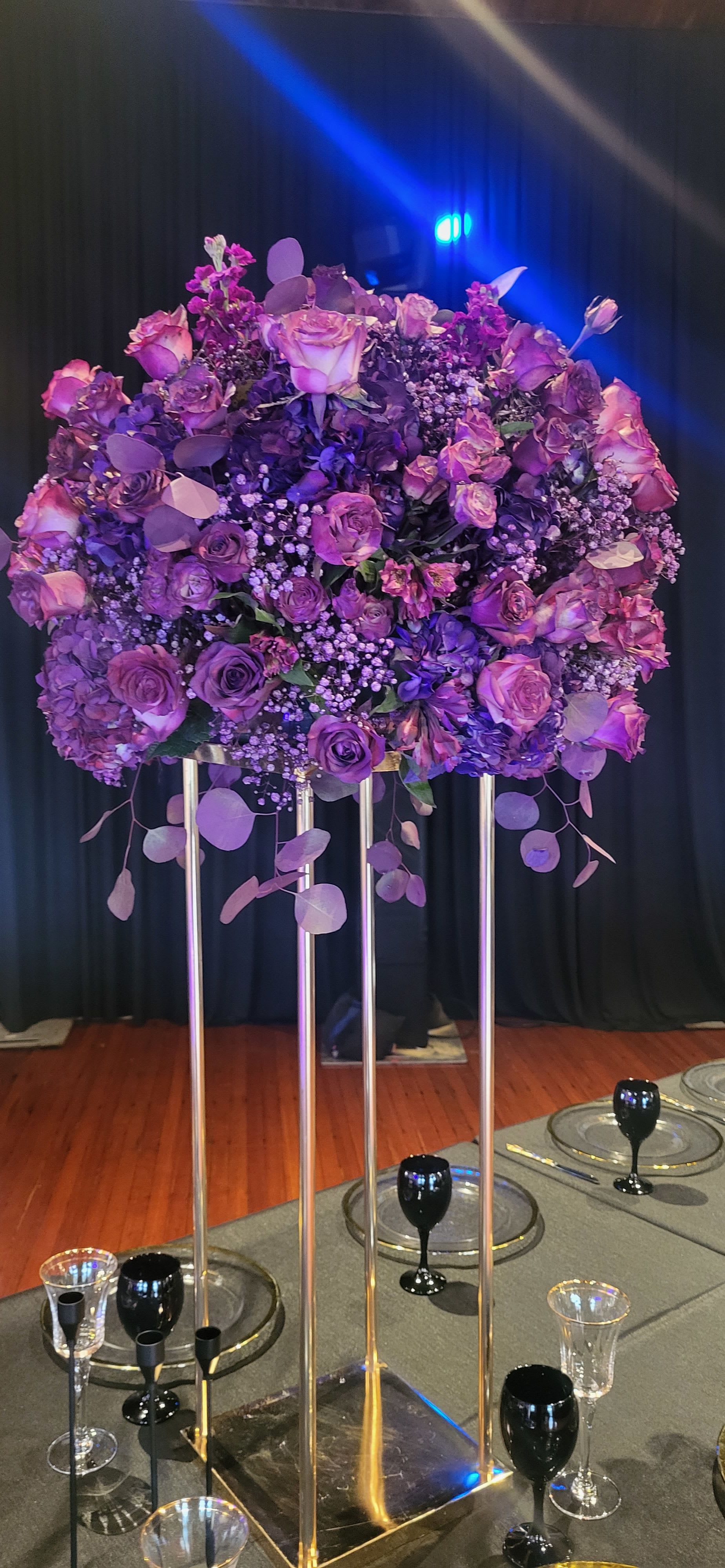 Purple center piece for special events no vase  - A lush, elevated centerpiece in rich purple tones-this flower arrangement for special events creates immediate drama and elegance for weddings, anniversaries, or upscale celebrations. The tall gold metal stand lifts a rounded mass of jewel-toned blooms so the person you're honoring feels seen from every seat; when paired with black linens or dark place settings the colors pop, making it an unforgettable focal point for a banquet or head table.  Densely packed roses, hydrangea-like clusters, carnations, and small filler flowers form the voluminous top, with round eucalyptus-style leaves trailing beneath to soften the silhouette. The palette mixes deep purple, soft lavender, bright pink and white accents for depth and contrast; the arrangement is designed without a vase for secure mounting on the included or venue-provided gold stand. Sized for long tables and high-impact moments, the overall look is formal and lush-perfect for couples, honored guests, or milestone celebrations.  Place these elevated centerpieces along banquet tables or at the ceremony entrance to set a cohesive, luxurious tone; they pair beautifully with black tablecloths, gold chargers, and mixed glassware like clear goblets and black wine glasses. Because the design is intended for event display, confirm stand and table dimensions with your venue when ordering to ensure the height and spread complement your seating plan.